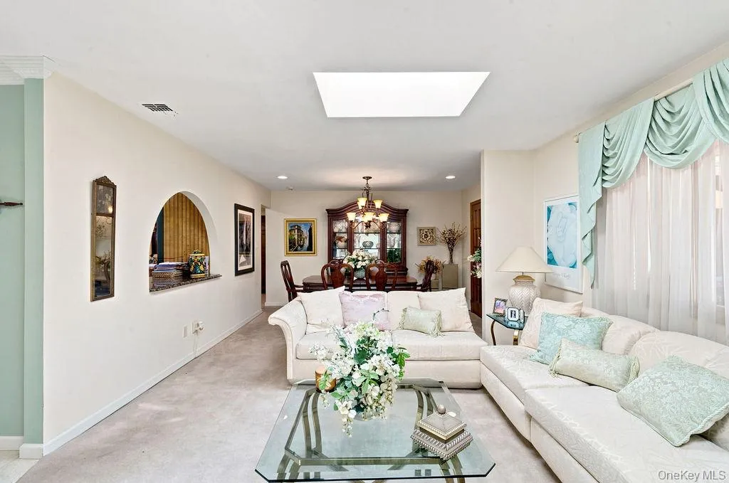 Recessed lighting and a rectangular skylight illuminate the living space, featuring wall-to-wall carpeting and an arched pass-through to a formal dining area with a chandelier Recessed lighting and a rectangular skylight illuminate the living space, featuring wall-to-wall carpeting and an arched pass-through to a formal dining area with a chandelier
