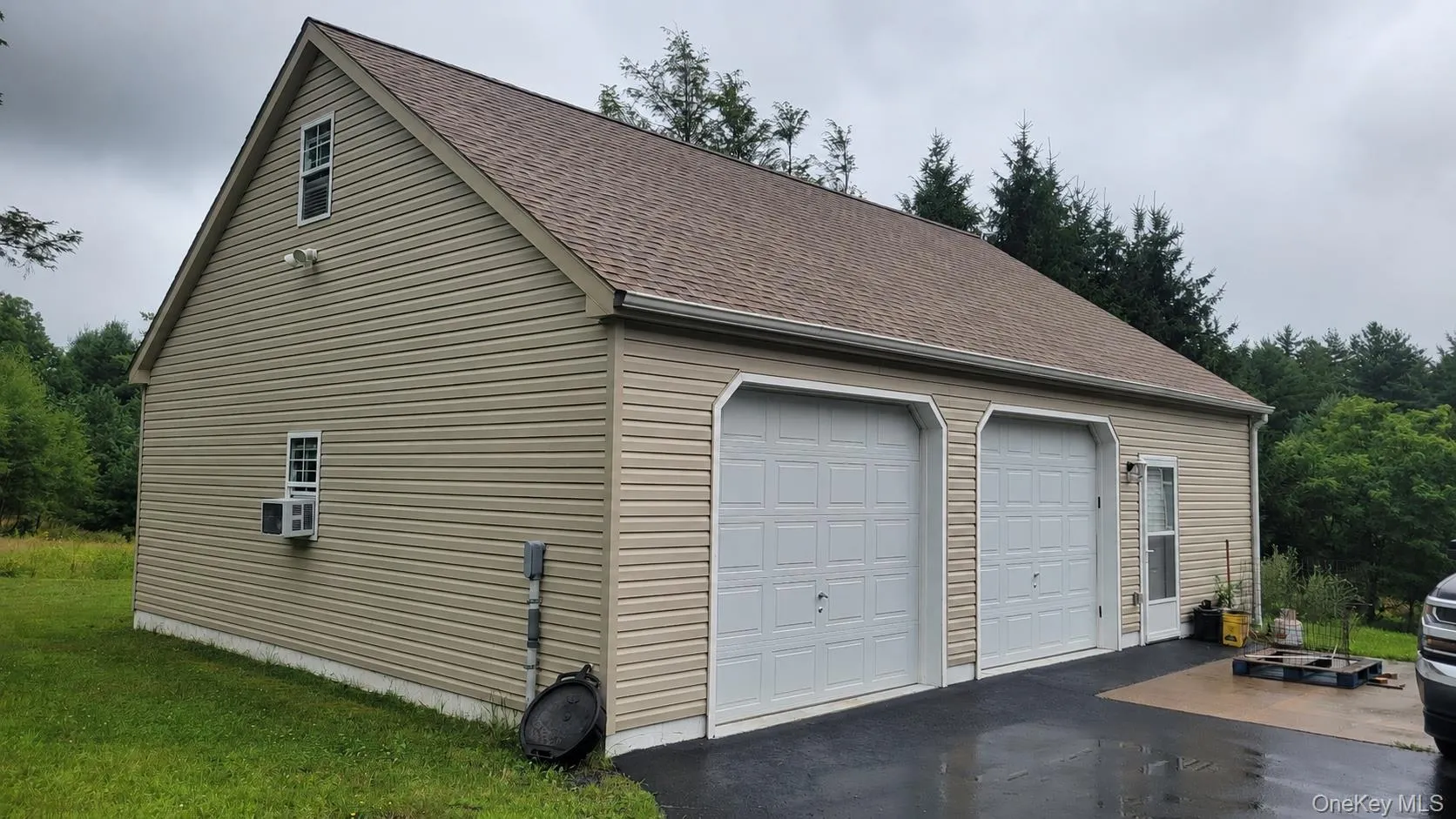 Two-story garage structure featuring tan horizontal siding, a brown shingle roof, two white panel garage doors, a side entry door, and multiple windows Two-story garage structure featuring tan horizontal siding, a brown shingle roof, two white panel garage doors, a side entry door, and multiple windows