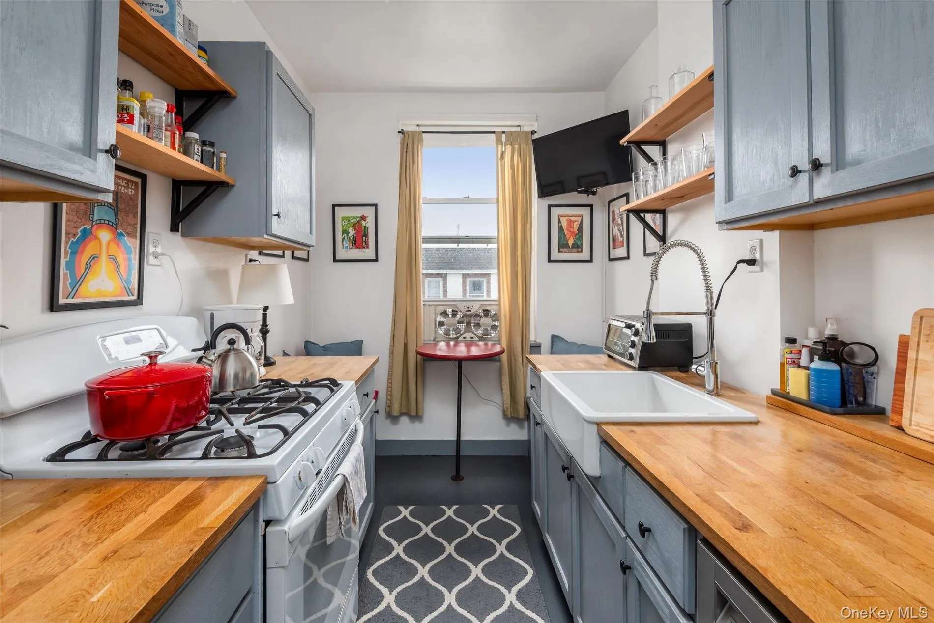 Kitchen featuring wood-finish countertops, blue cabinetry with dark hardware, a white farmhouse sink with a gooseneck faucet, a white gas range, and open shelving Kitchen featuring wood-finish countertops, blue cabinetry with dark hardware, a white farmhouse sink with a gooseneck faucet, a white gas range, and open shelving