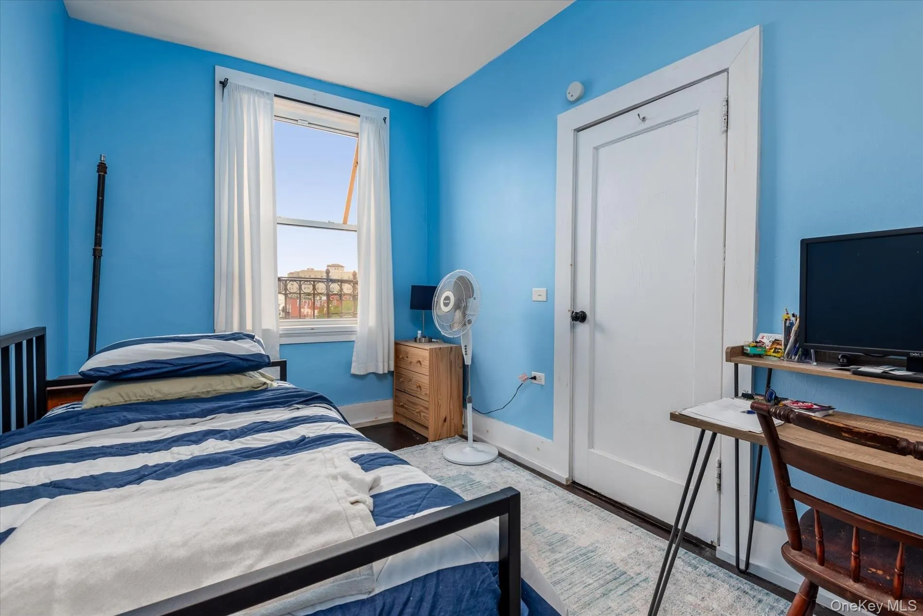 Bright room featuring a single window with white curtains, a white panel door, and dark wood-finish flooring Bright room featuring a single window with white curtains, a white panel door, and dark wood-finish flooring
