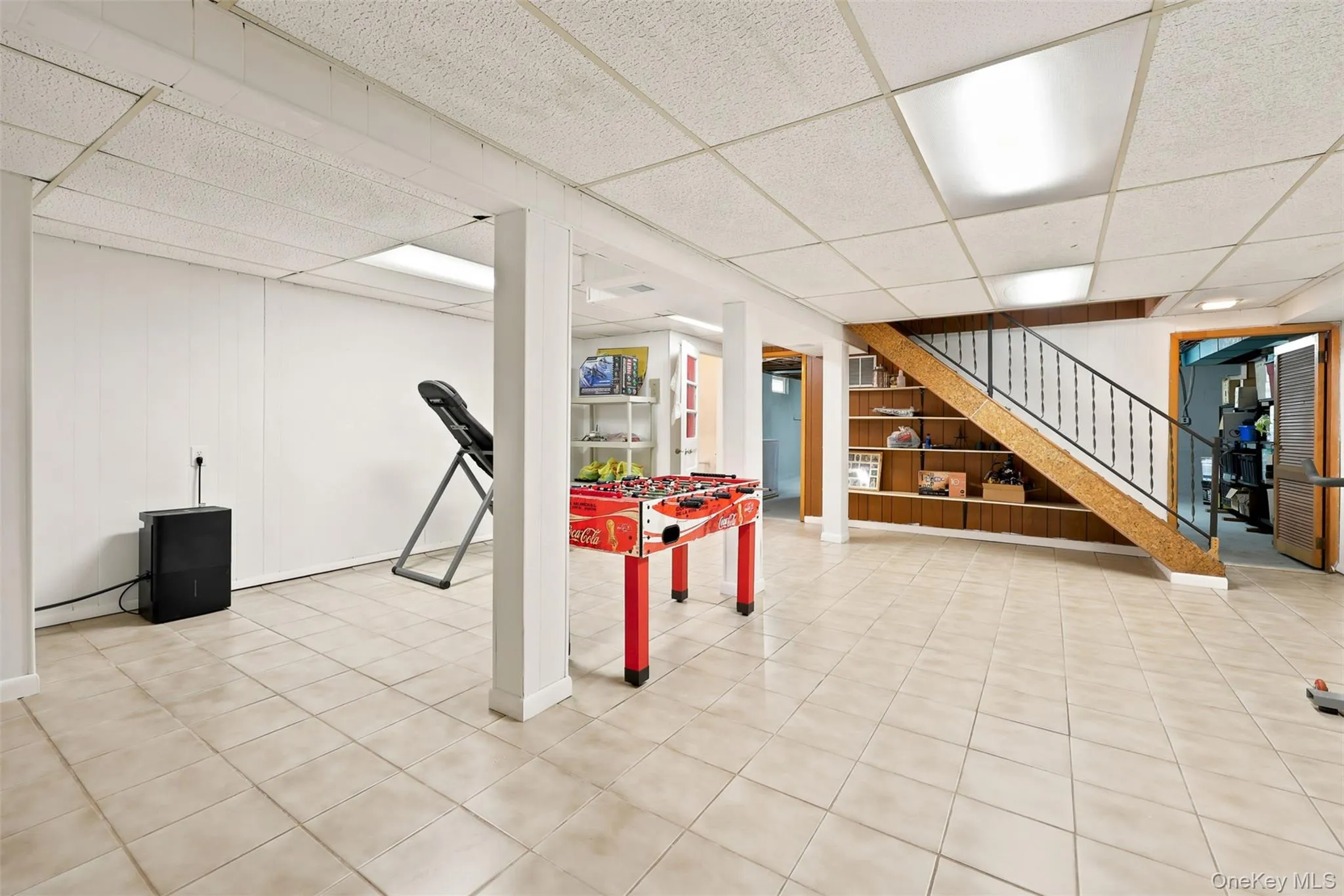 Spacious finished basement featuring light-toned tile flooring, white painted walls, and a drop ceiling with integrated fluorescent lighting Spacious finished basement featuring light-toned tile flooring, white painted walls, and a drop ceiling with integrated fluorescent lighting
