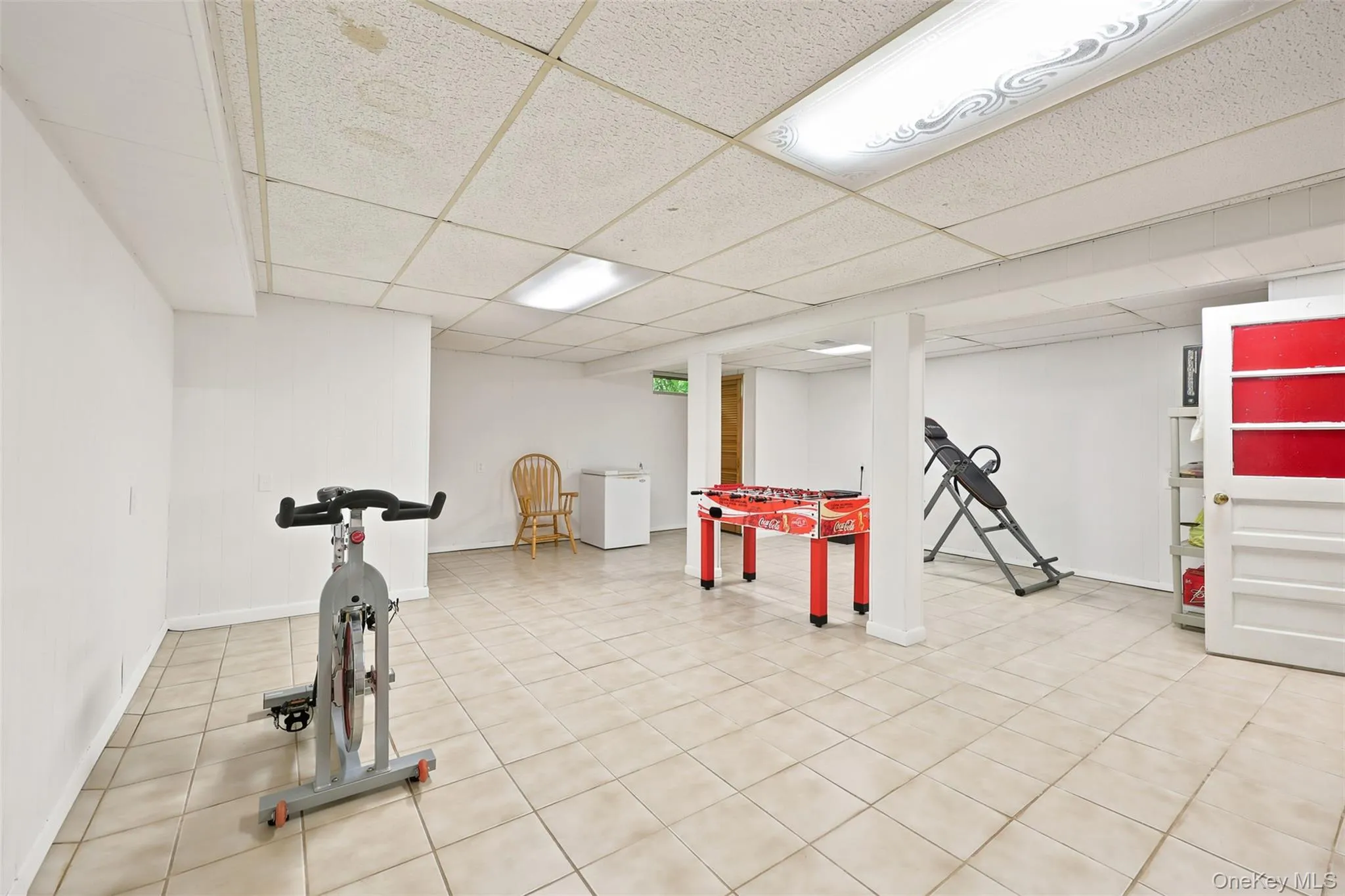 Spacious finished basement featuring light-toned tile flooring, white painted walls, and a drop ceiling with integrated fluorescent lighting Spacious finished basement featuring light-toned tile flooring, white painted walls, and a drop ceiling with integrated fluorescent lighting