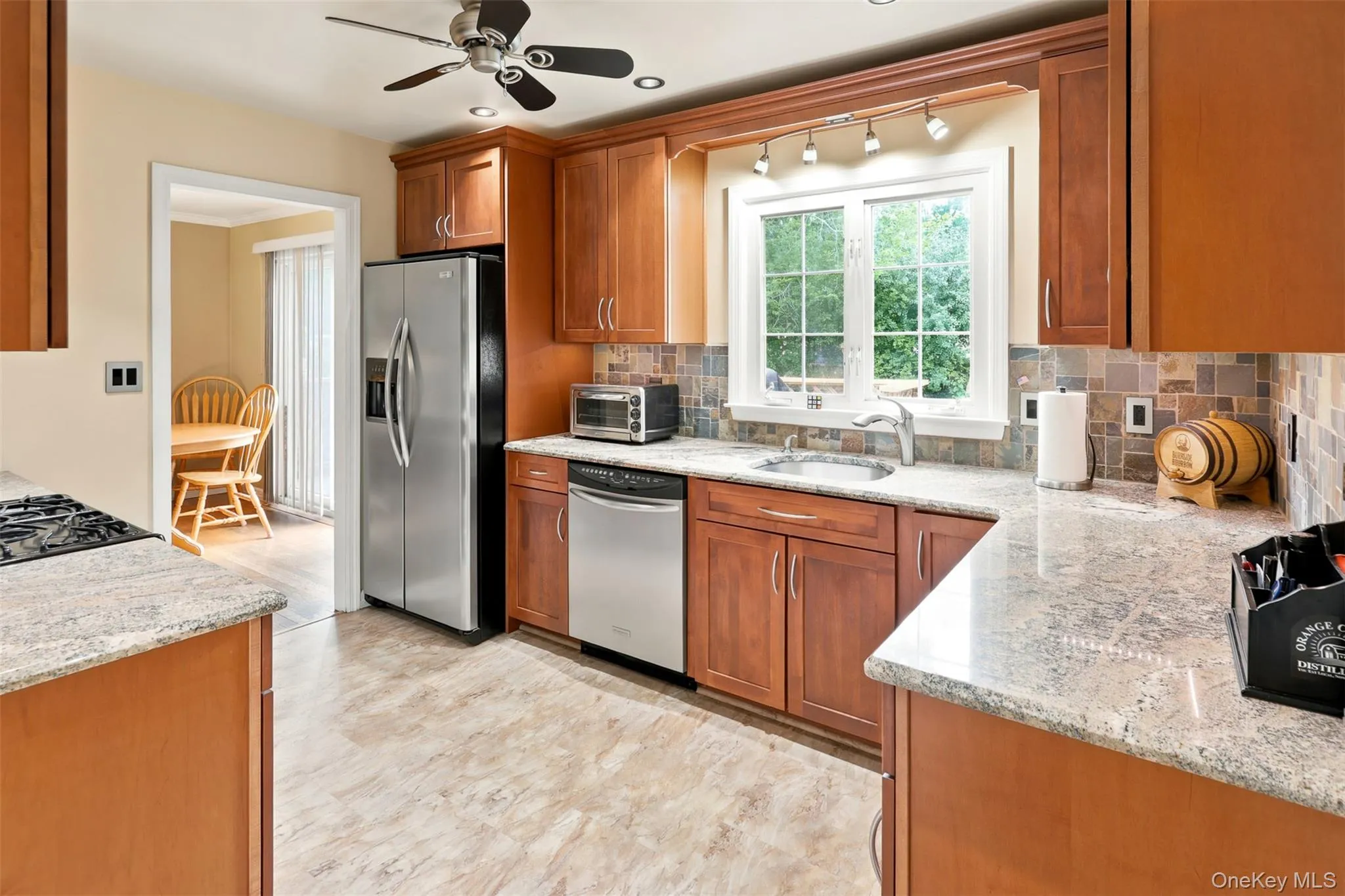Kitchen featuring wood cabinetry, light-toned countertops, a multi-tone tile backsplash, stainless steel appliances, and wood-finish flooring Kitchen featuring wood cabinetry, light-toned countertops, a multi-tone tile backsplash, stainless steel appliances, and wood-finish flooring
