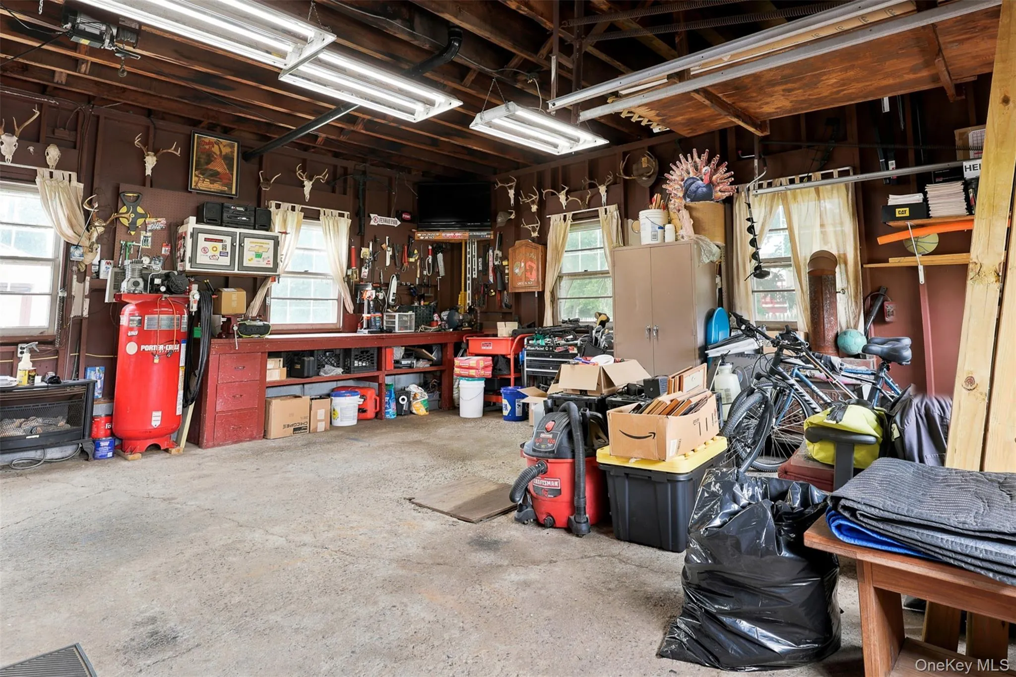 Spacious garage featuring a concrete floor, exposed wood beam ceiling, multiple fluorescent light fixtures, and two windows providing natural light Spacious garage featuring a concrete floor, exposed wood beam ceiling, multiple fluorescent light fixtures, and two windows providing natural light