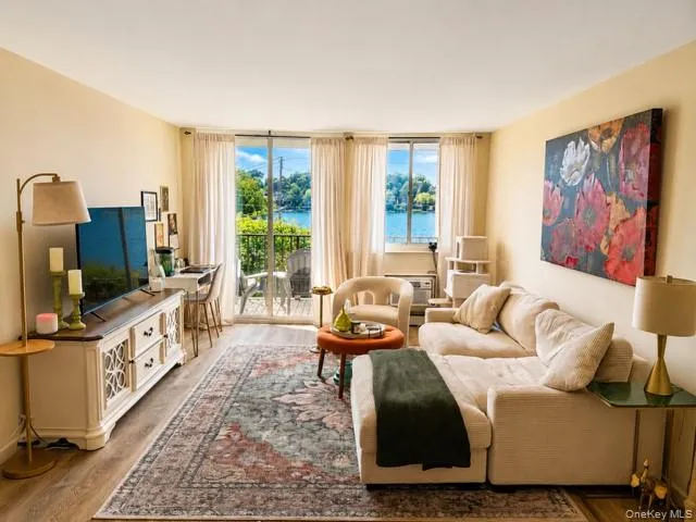 Bright, light-filled living room showcasing expansive glass doors and windows with water views, neutral upholstered seating, hardwood-finish flooring, a patterned area rug, built-in media console with storage cabinetry, brass floor lamp, and contemporary artwork Bright, light-filled living room showcasing expansive glass doors and windows with water views, neutral upholstered seating, hardwood-finish flooring, a patterned area rug, built-in media console with storage cabinetry, brass floor lamp, and contemporary artwork