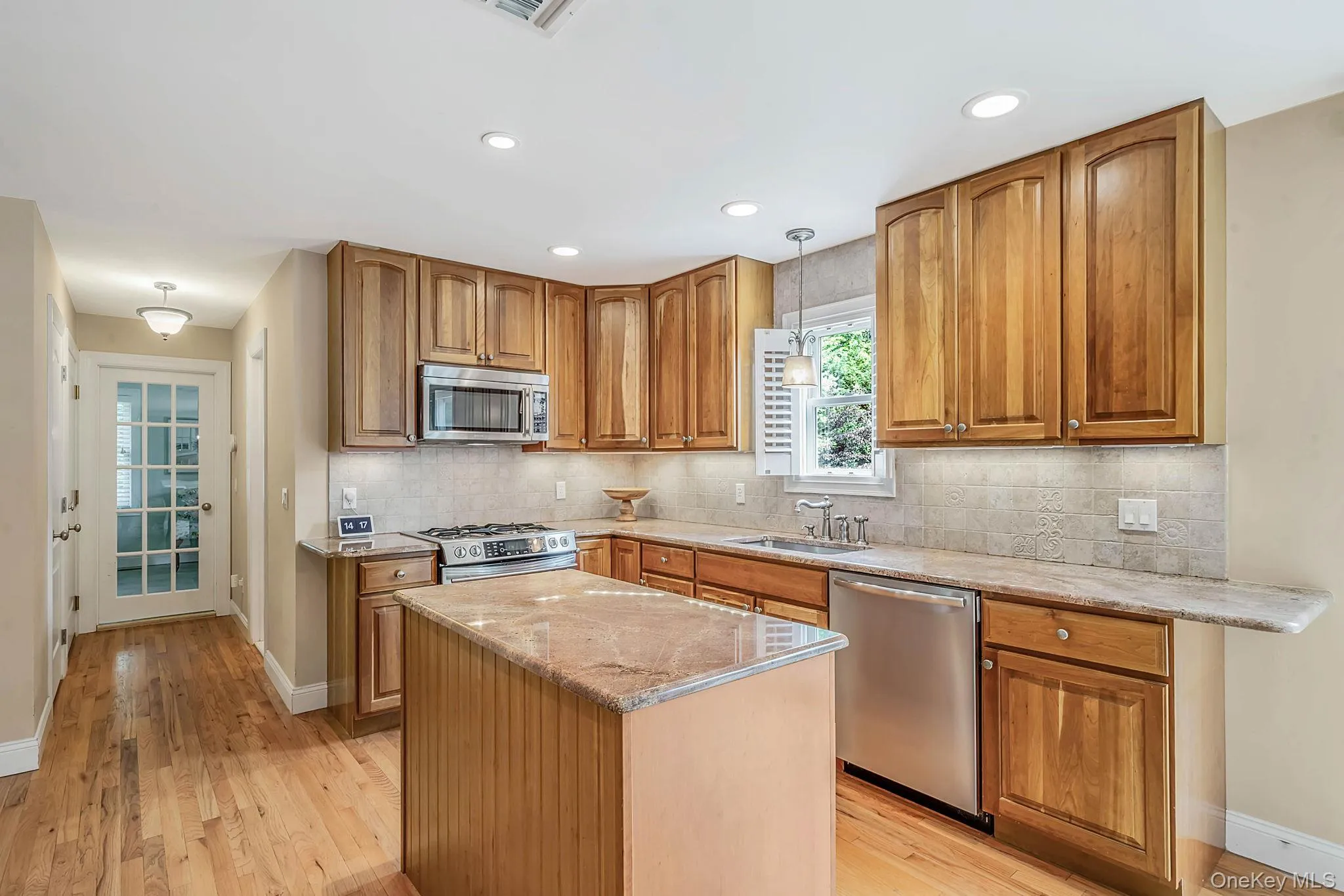 Kitchen featuring wood cabinetry, stone countertops, stone tile backsplash, stainless steel appliances, and wood-finish flooring Kitchen featuring wood cabinetry, stone countertops, stone tile backsplash, stainless steel appliances, and wood-finish flooring
