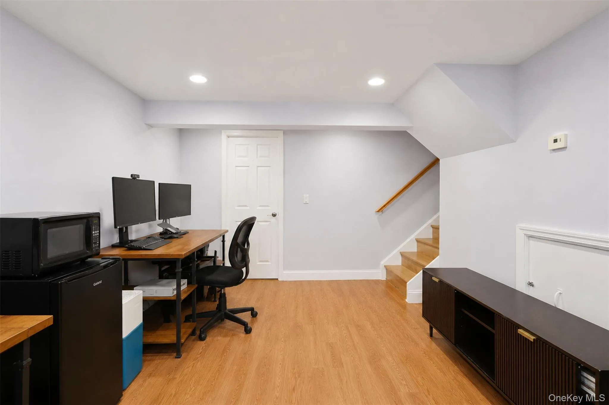 Versatile open space featuring wood-finish flooring, recessed lighting, a built-in staircase with wood handrail, and a white paneled door Versatile open space featuring wood-finish flooring, recessed lighting, a built-in staircase with wood handrail, and a white paneled door