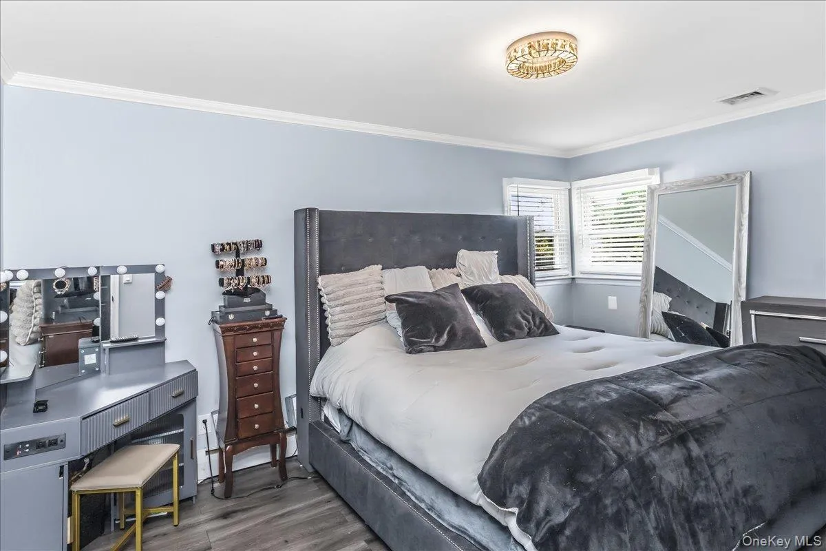 Spacious room featuring light blue walls, crown molding, a decorative flush-mount ceiling fixture, two windows with blinds, and wood-finish flooring Spacious room featuring light blue walls, crown molding, a decorative flush-mount ceiling fixture, two windows with blinds, and wood-finish flooring
