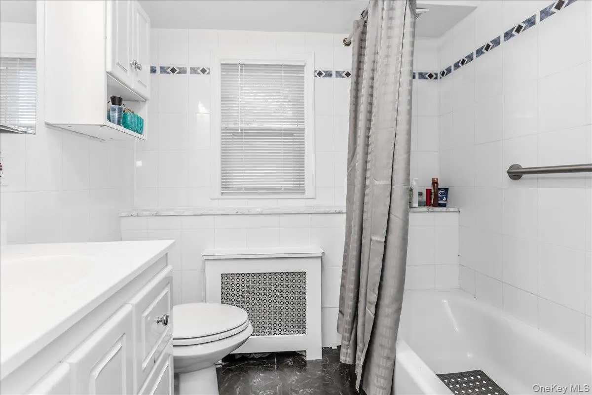 Bathroom featuring white wall tiling with decorative accent borders, a built-in bathtub with shower, a window with blinds, and a vanity with integrated sink Bathroom featuring white wall tiling with decorative accent borders, a built-in bathtub with shower, a window with blinds, and a vanity with integrated sink