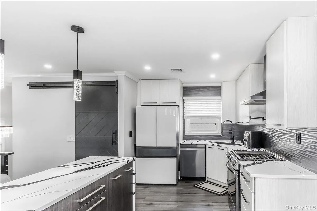 Modern kitchen featuring white cabinetry, a large center island with a white and black veined countertop, stainless steel appliances, wood-finish flooring, and a barn door entry Modern kitchen featuring white cabinetry, a large center island with a white and black veined countertop, stainless steel appliances, wood-finish flooring, and a barn door entry