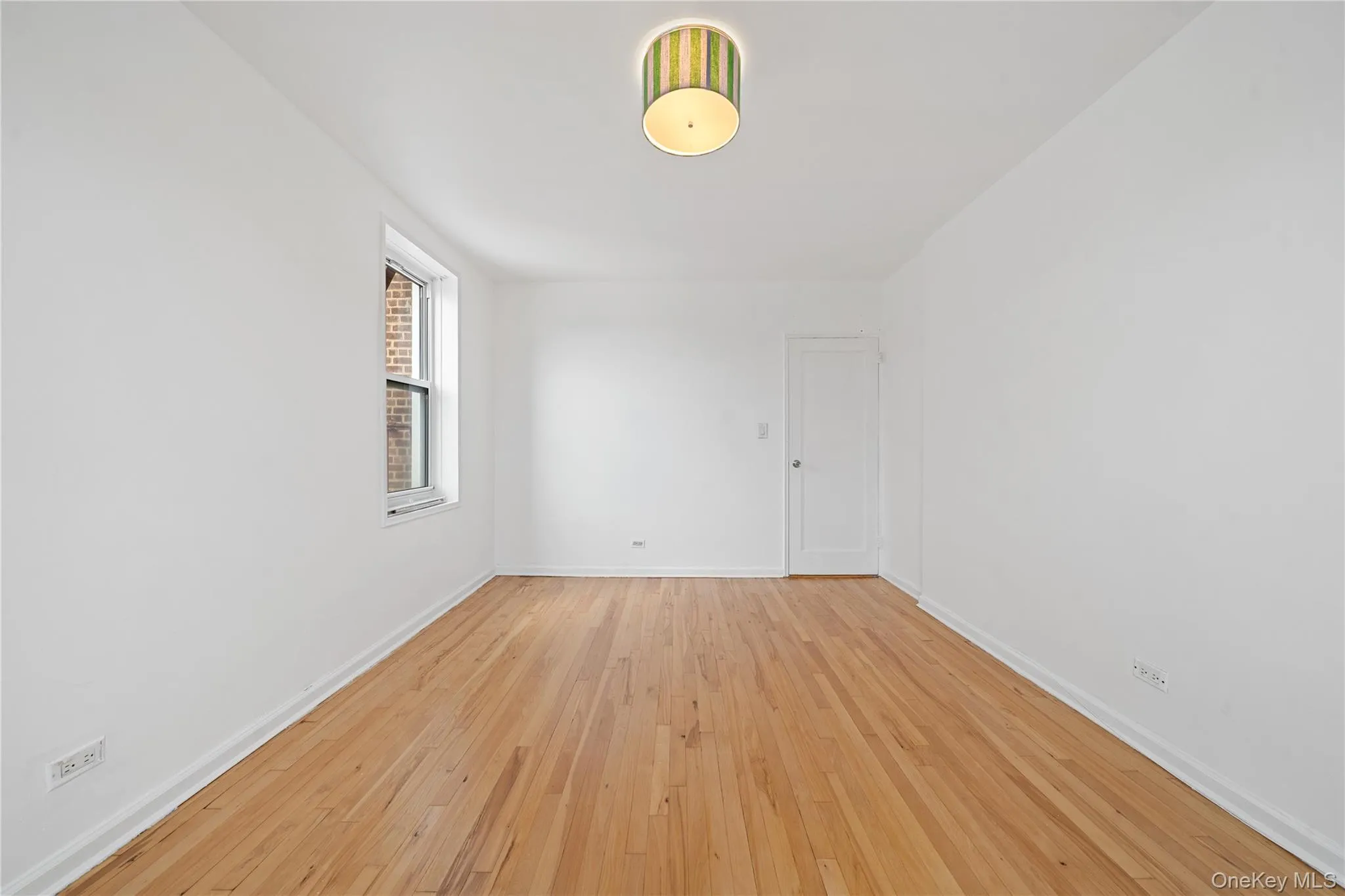 Hardwood flooring, white walls, single window, overhead flush mount light fixture, and a single paneled door Hardwood flooring, white walls, single window, overhead flush mount light fixture, and a single paneled door