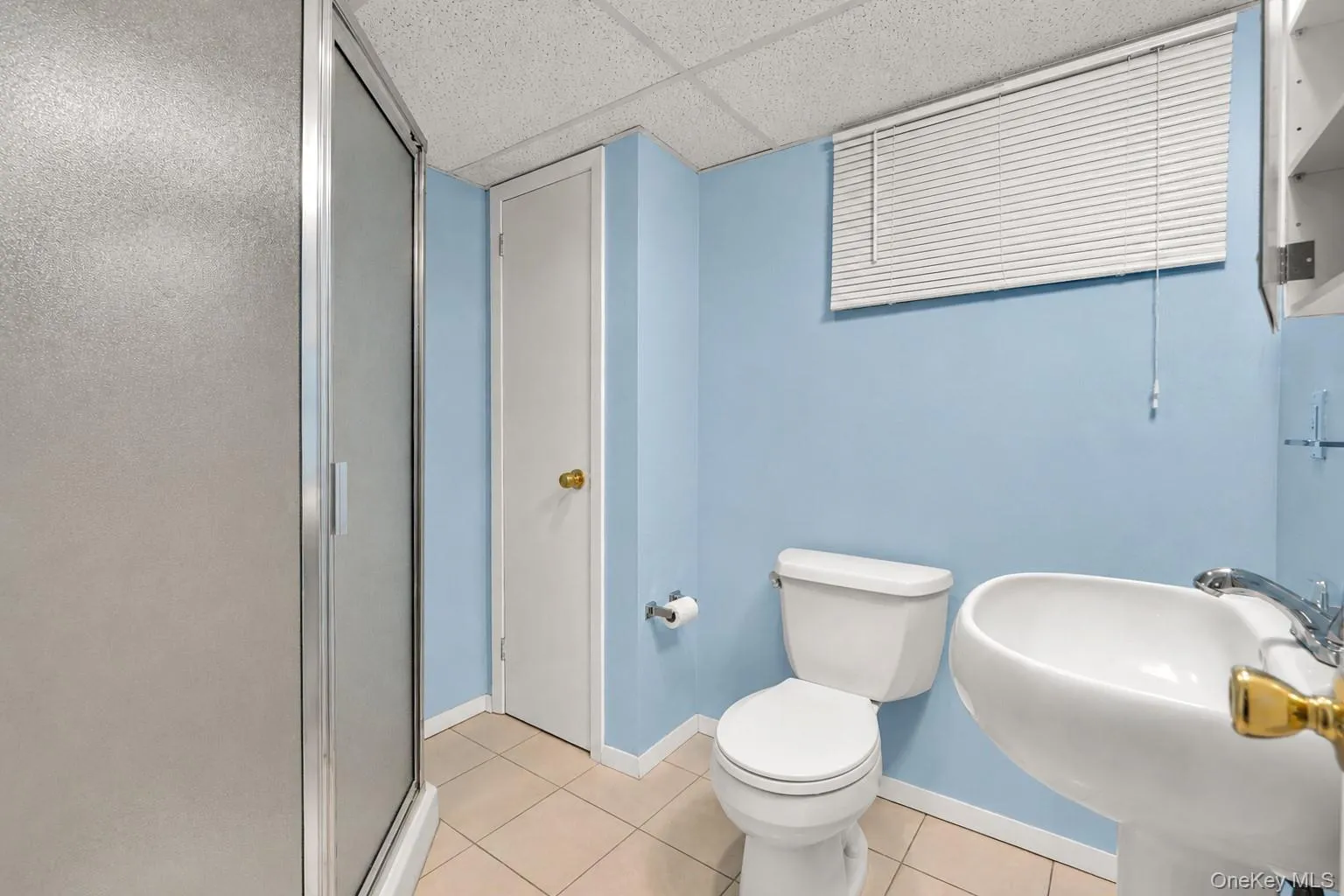 Bathroom in the basement featuring a glass-enclosed shower, pedestal sink with chrome faucet, toilet, and light-colored tile flooring Bathroom in the basement featuring a glass-enclosed shower, pedestal sink with chrome faucet, toilet, and light-colored tile flooring