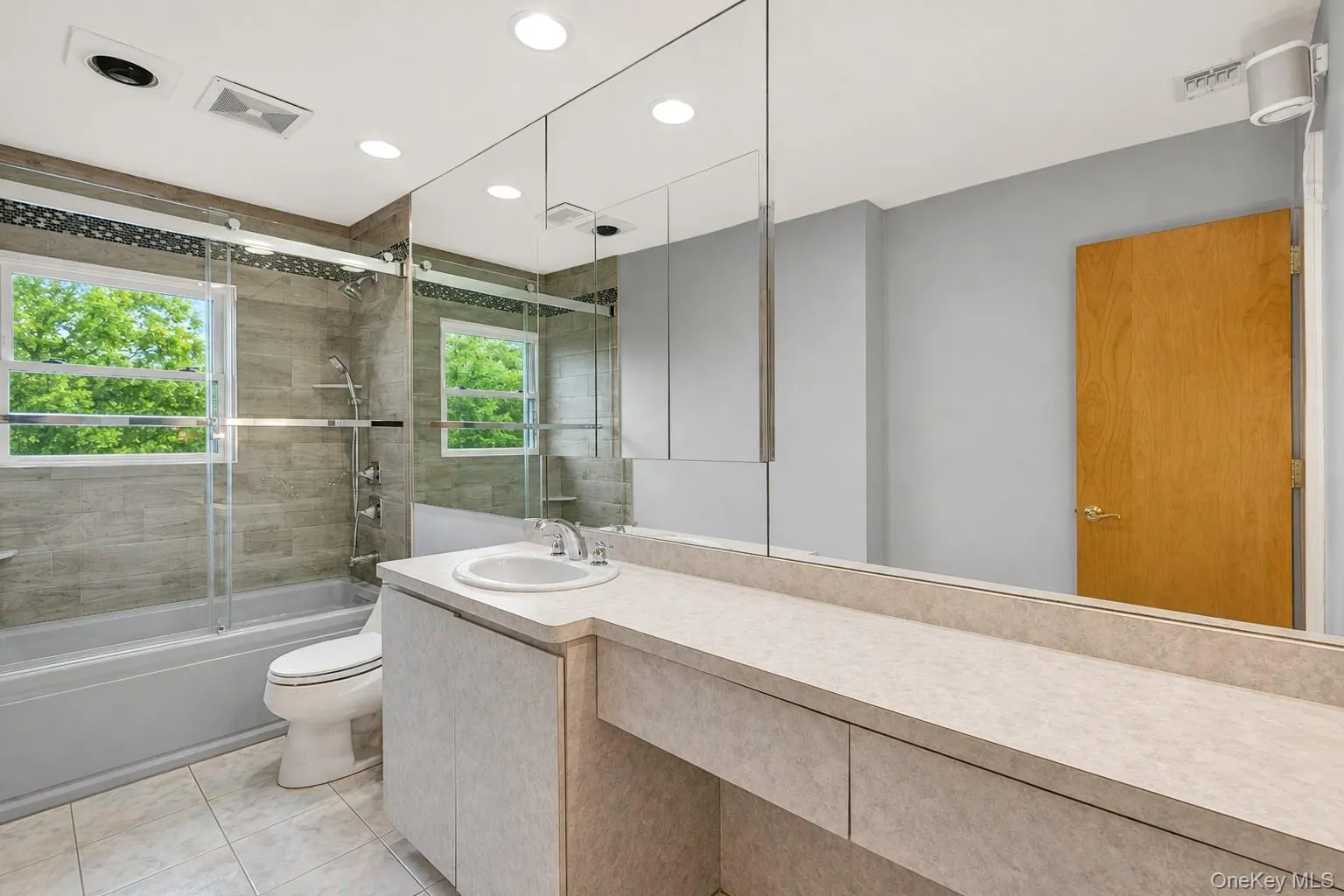 Bathroom featuring a large vanity with a single basin sink, a full-wall mirror, recessed lighting, a combination tub and shower with glass enclosure, and a tiled floor Bathroom featuring a large vanity with a single basin sink, a full-wall mirror, recessed lighting, a combination tub and shower with glass enclosure, and a tiled floor