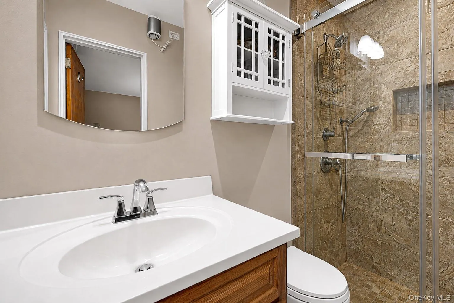 Bathroom featuring a wood-finish vanity with an integrated sink and chrome faucet, a curved vanity mirror, and a glass-enclosed shower with stone-look wall tiling Bathroom featuring a wood-finish vanity with an integrated sink and chrome faucet, a curved vanity mirror, and a glass-enclosed shower with stone-look wall tiling