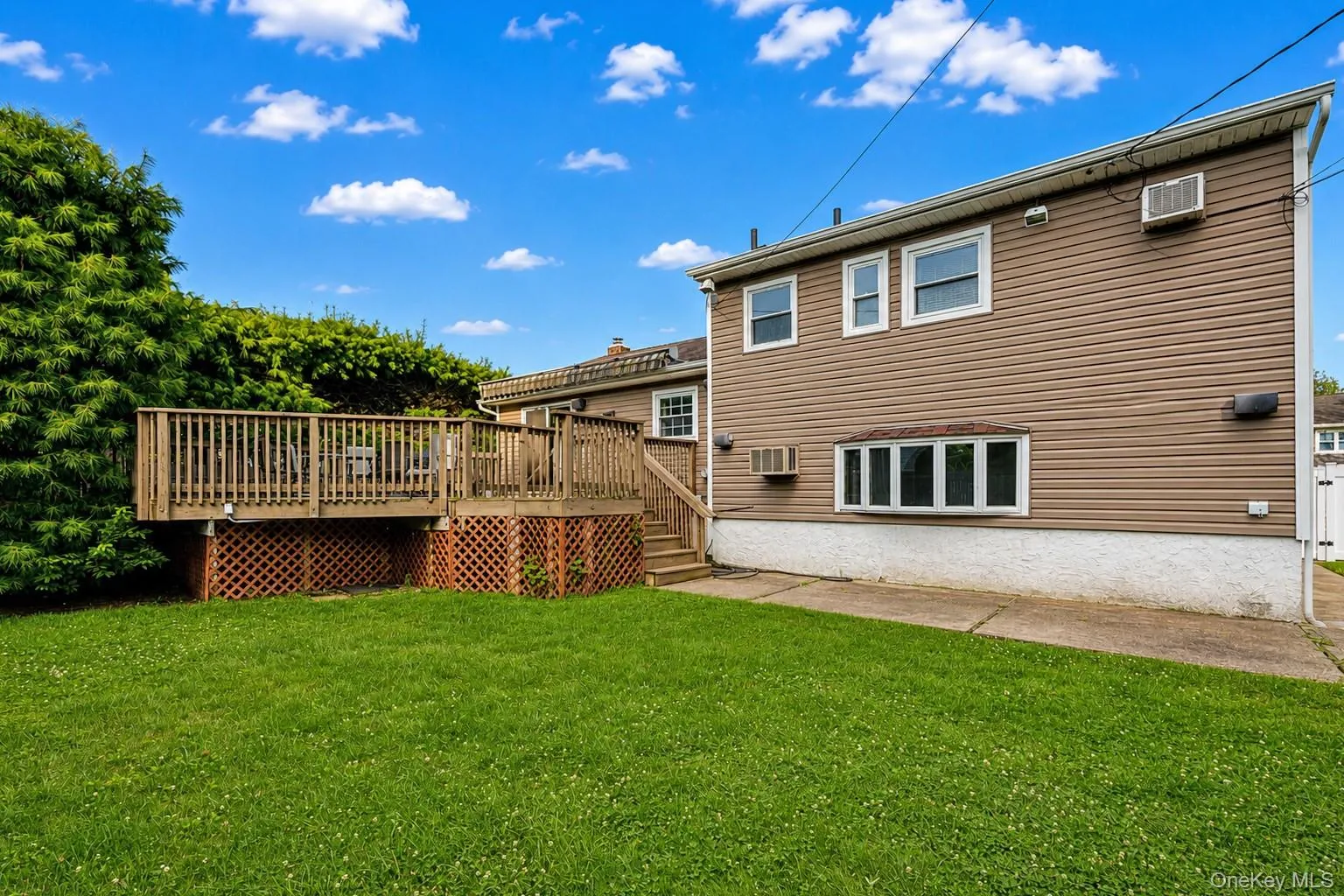 Expansive backyard featuring a large wooden deck with lattice skirting, a manicured lawn, mature perimeter landscaping, and a concrete patio area Expansive backyard featuring a large wooden deck with lattice skirting, a manicured lawn, mature perimeter landscaping, and a concrete patio area
