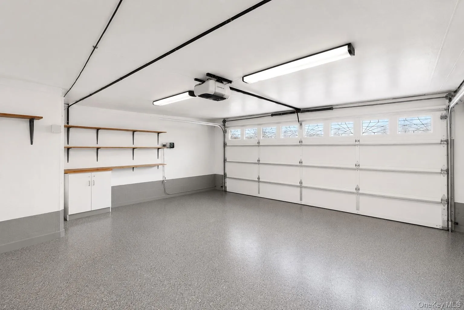 Spacious 2 car garage featuring epoxy-coated concrete flooring, white paneled overhead door with transom windows, wall-mounted cabinetry with wood-finish shelving, integrated ceiling-mounted lighting system, and ample natural light from clerestory windows Spacious 2 car garage featuring epoxy-coated concrete flooring, white paneled overhead door with transom windows, wall-mounted cabinetry with wood-finish shelving, integrated ceiling-mounted lighting system, and ample natural light from clerestory windows
