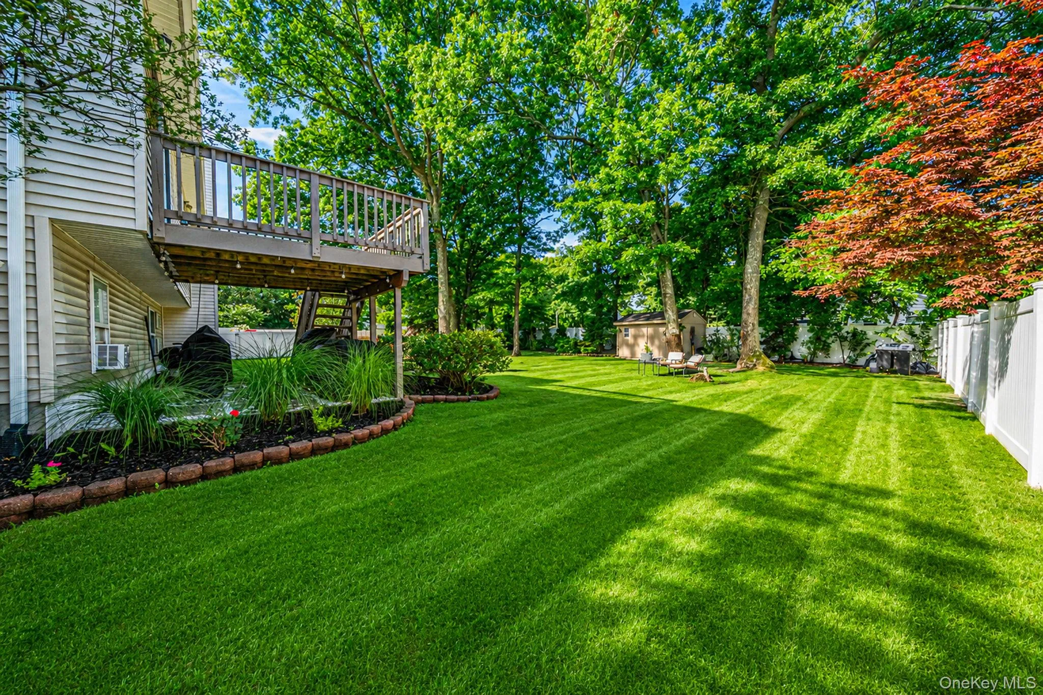 Expansive rear yard featuring a large wooden deck, lower patio area, mature trees, a storage shed, and a white privacy fence Expansive rear yard featuring a large wooden deck, lower patio area, mature trees, a storage shed, and a white privacy fence