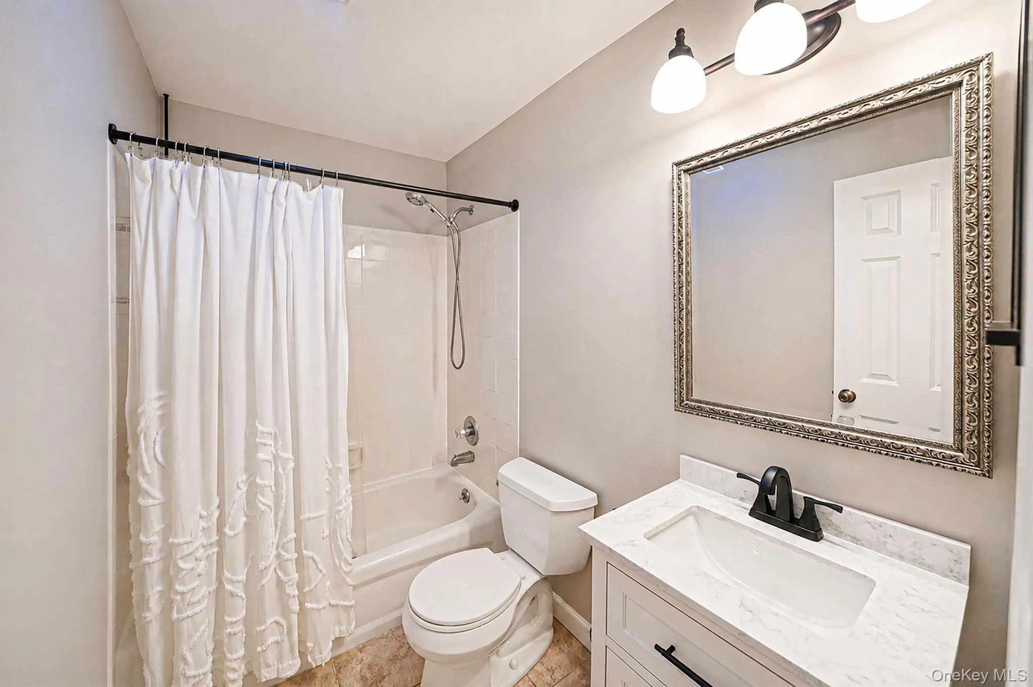 Bathroom featuring a white vanity with a white countertop and rectangular sink, a decorative framed mirror, a three-light vanity fixture, and a shower/tub combination Bathroom featuring a white vanity with a white countertop and rectangular sink, a decorative framed mirror, a three-light vanity fixture, and a shower/tub combination