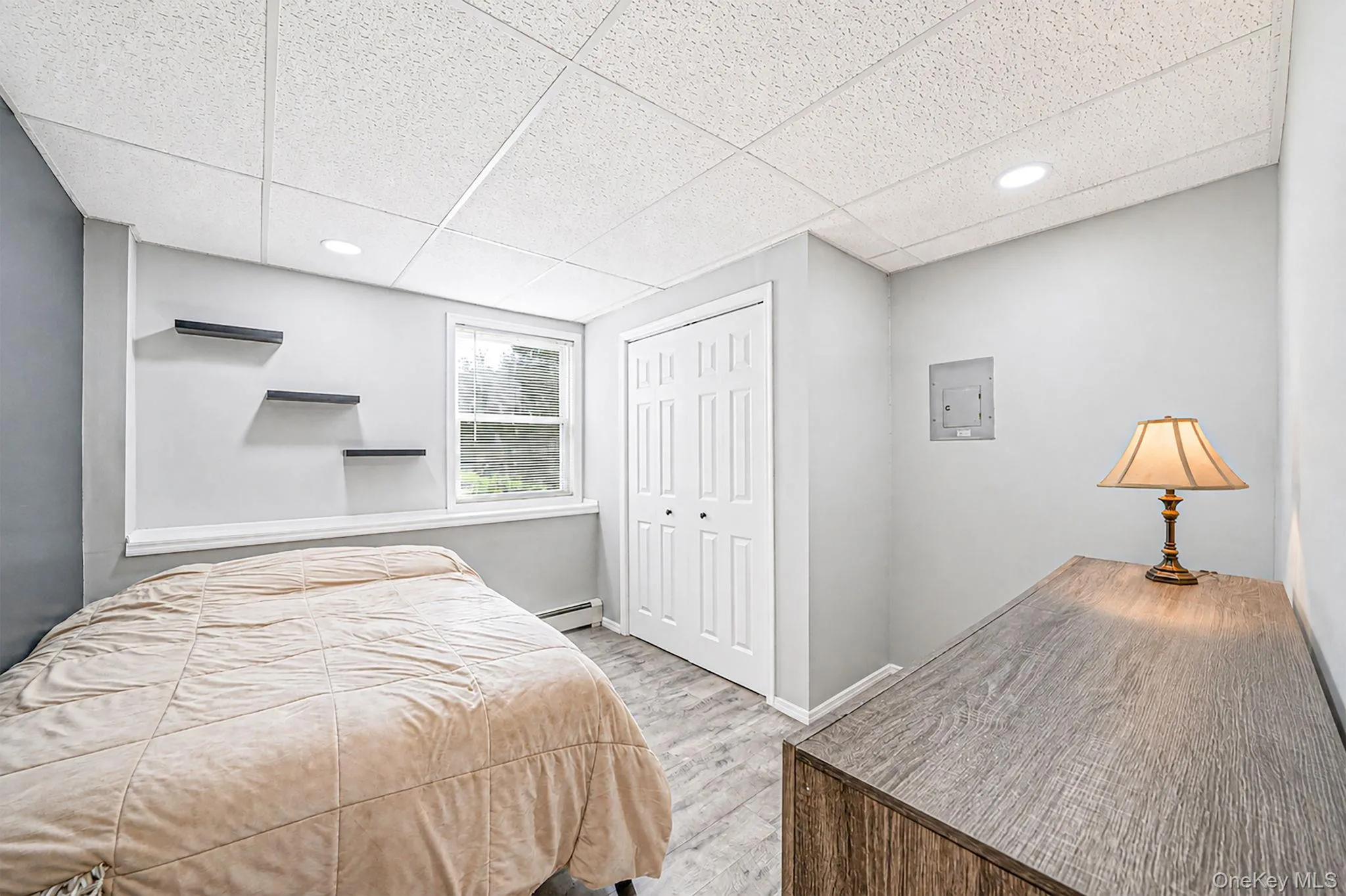 Bedroom featuring light gray walls, wood-finish flooring, a white bi-fold closet door, a single window with white trim, and a white grid-patterned ceiling with recessed lighting Bedroom featuring light gray walls, wood-finish flooring, a white bi-fold closet door, a single window with white trim, and a white grid-patterned ceiling with recessed lighting
