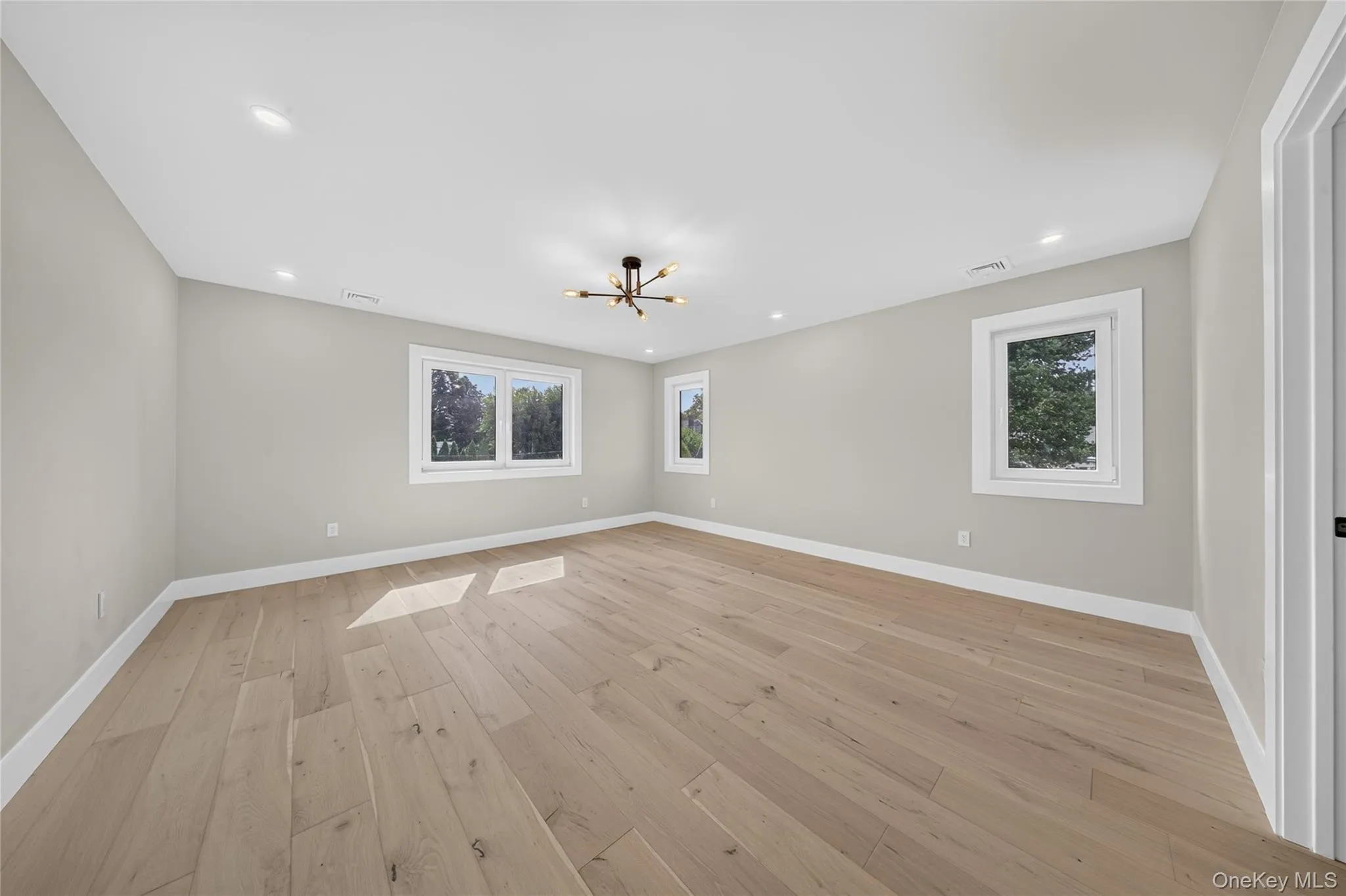 Spacious room with light wood-finish flooring, recessed lighting, and a modern ceiling fixture Spacious room with light wood-finish flooring, recessed lighting, and a modern ceiling fixture