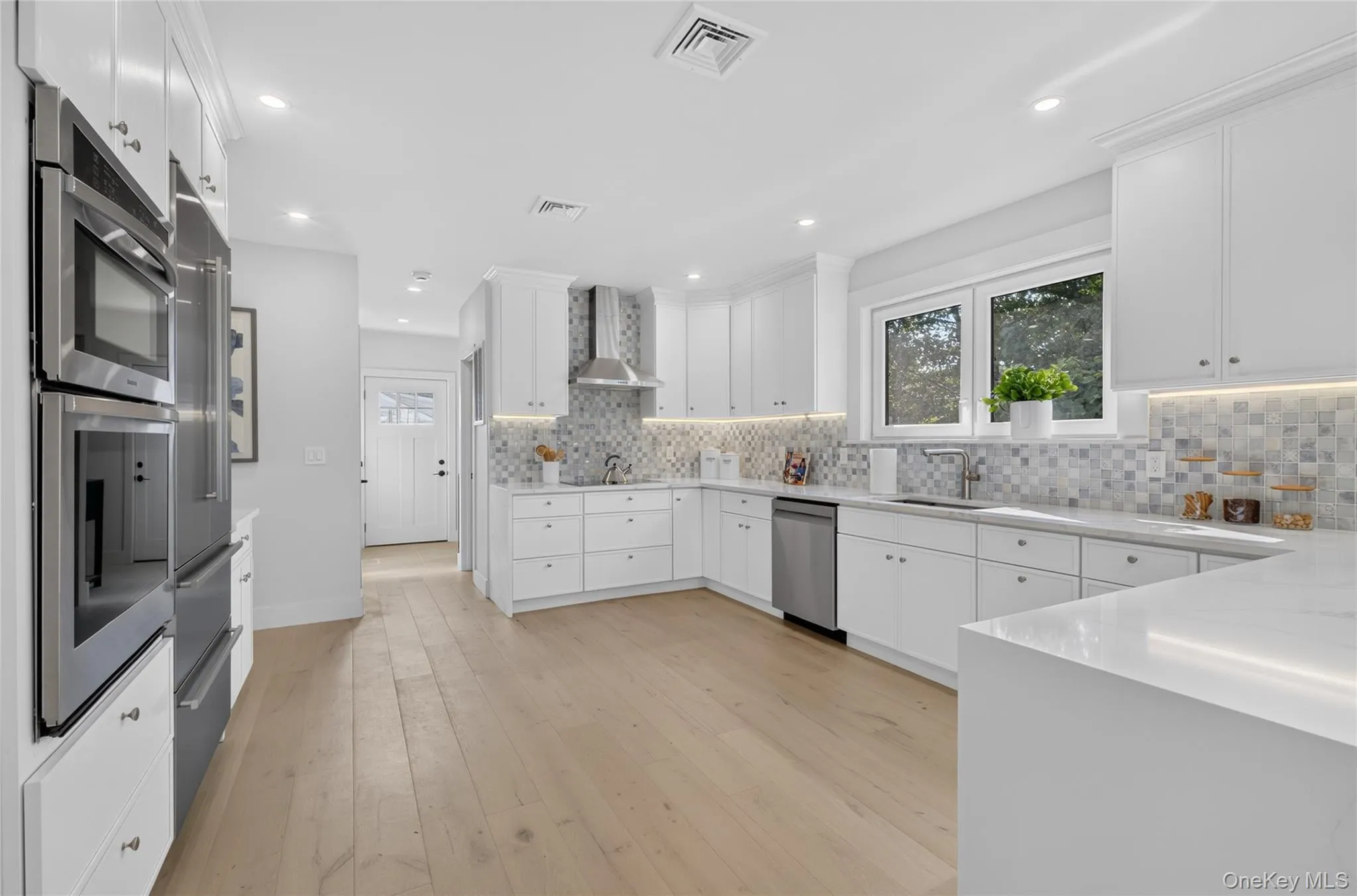 Kitchen featuring wood-finish flooring, white cabinetry, integrated stainless steel appliances, a mosaic tile backsplash, and light-toned countertops Kitchen featuring wood-finish flooring, white cabinetry, integrated stainless steel appliances, a mosaic tile backsplash, and light-toned countertops
