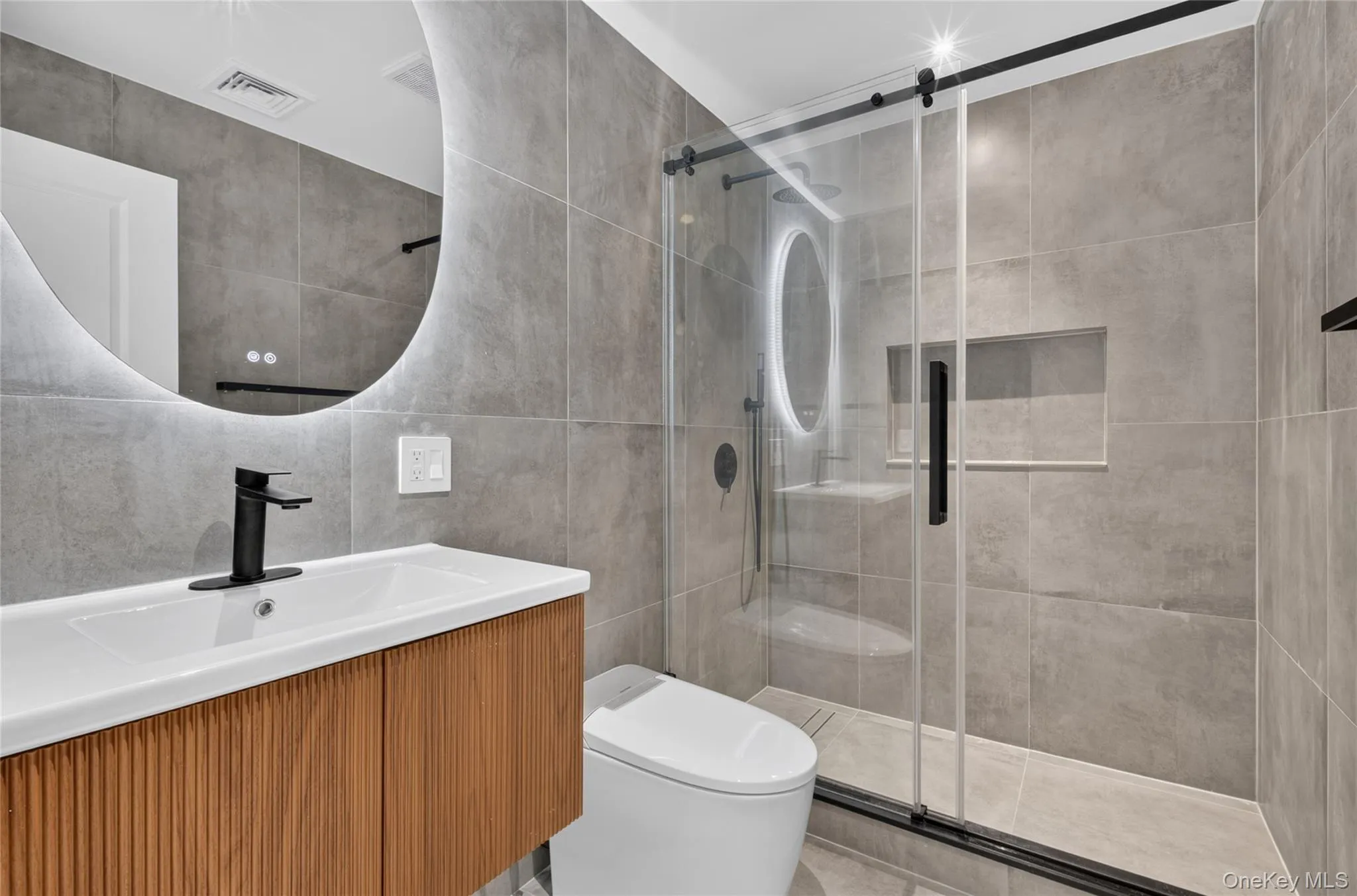 Modern bathroom featuring a wood-finish vanity, integrated sink, and an illuminated circular mirror Modern bathroom featuring a wood-finish vanity, integrated sink, and an illuminated circular mirror