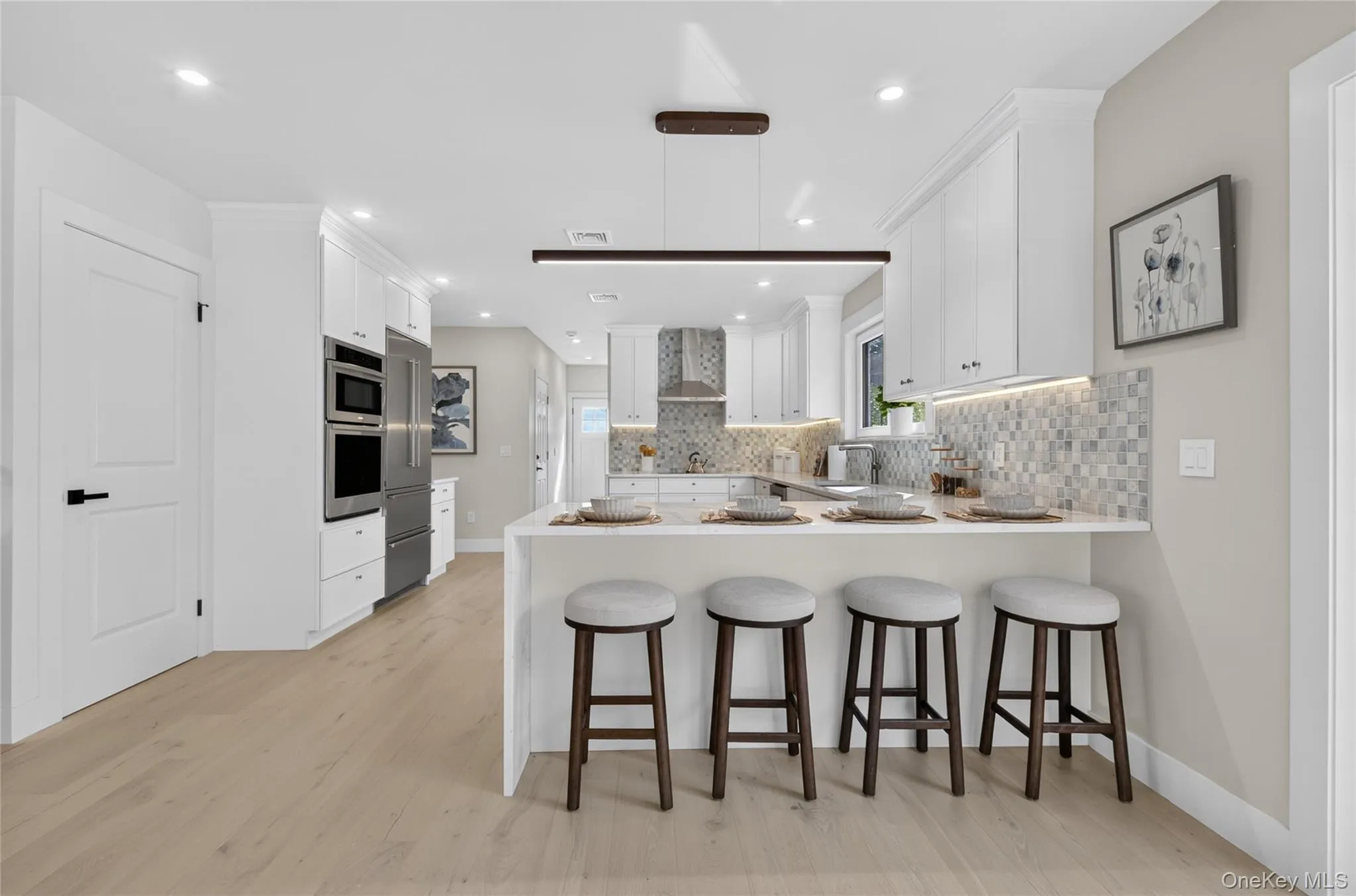 Modern kitchen featuring white cabinetry, light wood-finish flooring, stainless steel appliances, a breakfast bar, and a patterned tile backsplash Modern kitchen featuring white cabinetry, light wood-finish flooring, stainless steel appliances, a breakfast bar, and a patterned tile backsplash