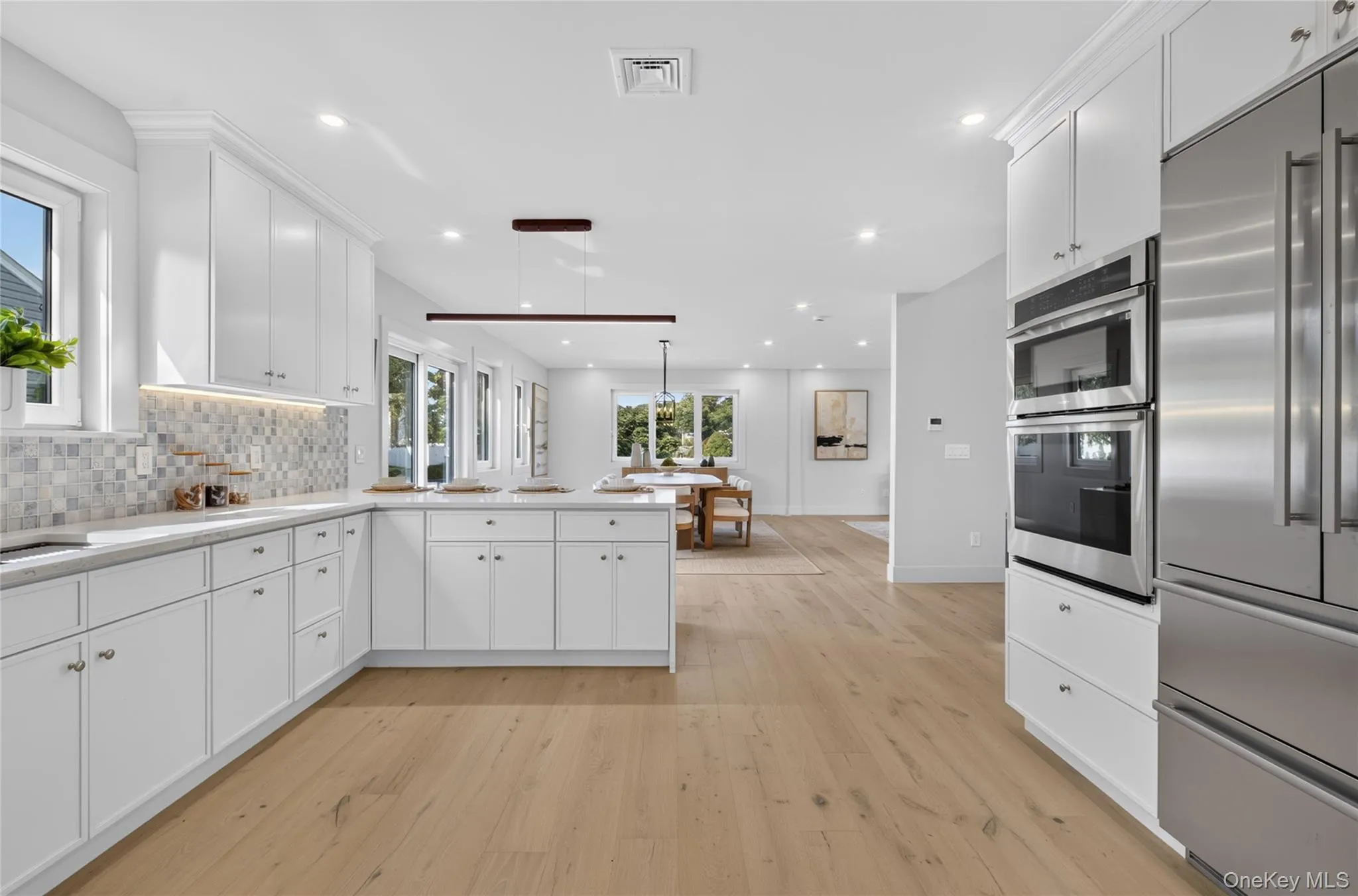Open-concept kitchen featuring extensive white cabinetry, light wood-finish flooring, stainless steel appliances, and a mosaic tile backsplash Open-concept kitchen featuring extensive white cabinetry, light wood-finish flooring, stainless steel appliances, and a mosaic tile backsplash