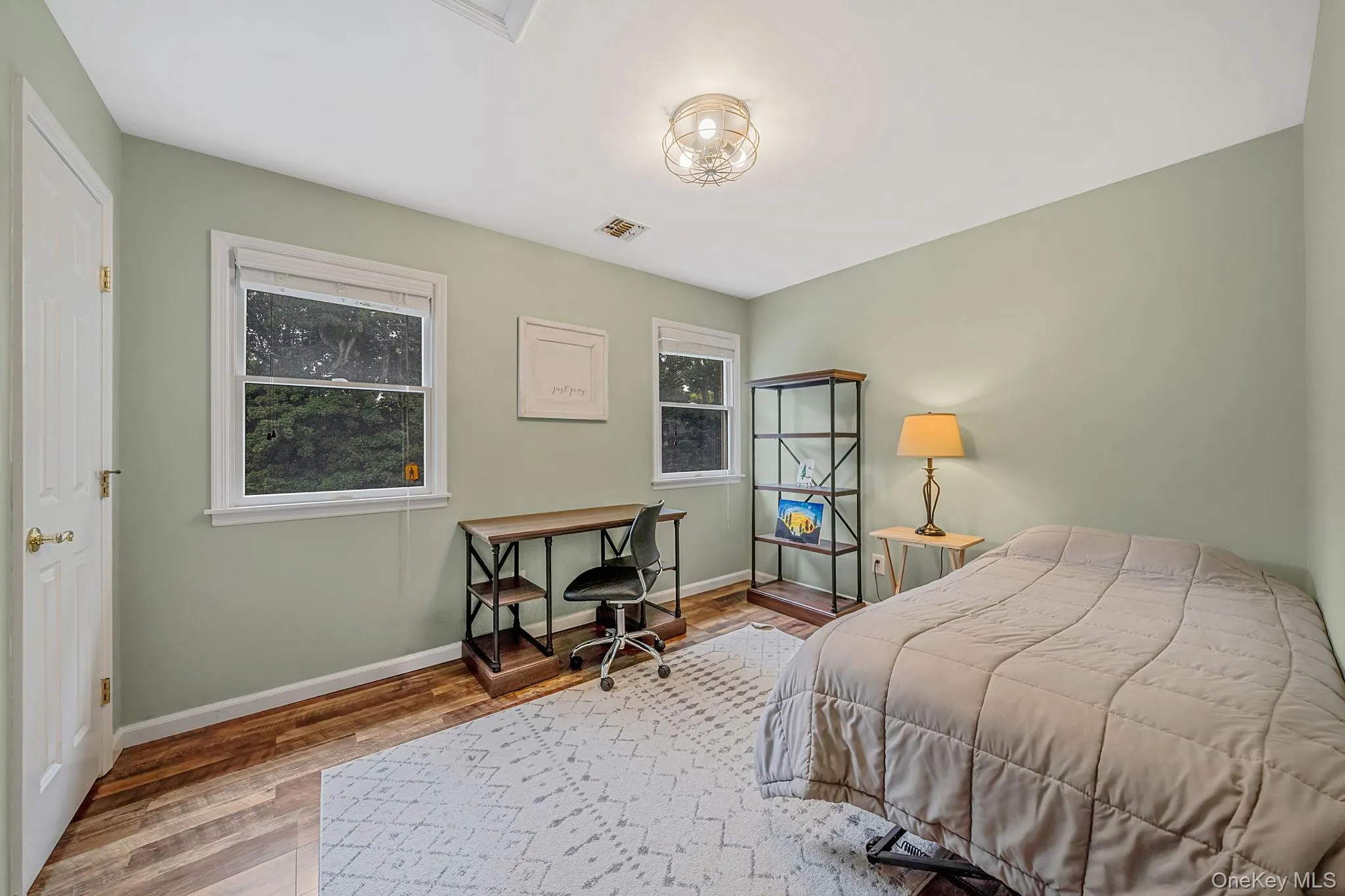 Bright secondary bedroom featuring soft sage-green walls, hardwood flooring, and generous natural light from dual windows Bright secondary bedroom featuring soft sage-green walls, hardwood flooring, and generous natural light from dual windows