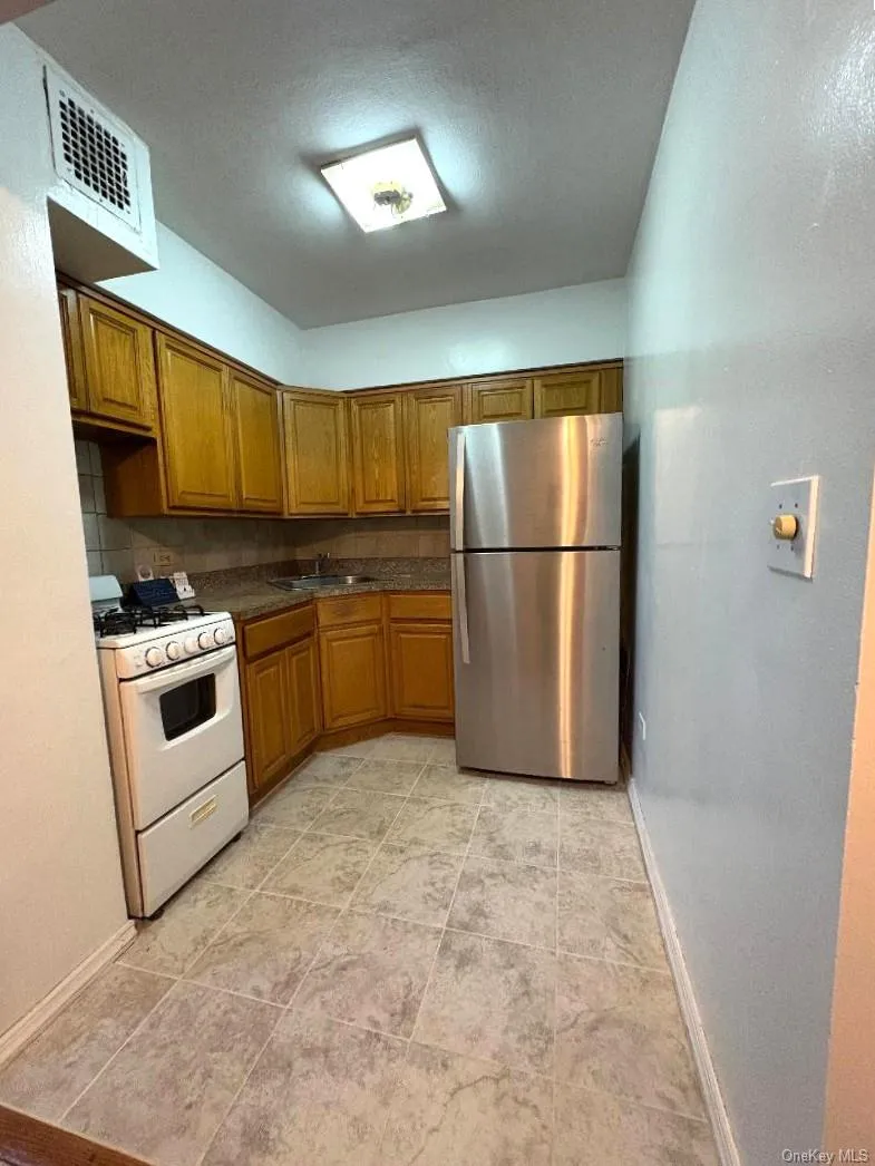 Kitchen featuring white gas range oven, freestanding refrigerator, brown cabinetry, tasteful backsplash, and light tile patterned floors Kitchen featuring white gas range oven, freestanding refrigerator, brown cabinetry, tasteful backsplash, and light tile patterned floors