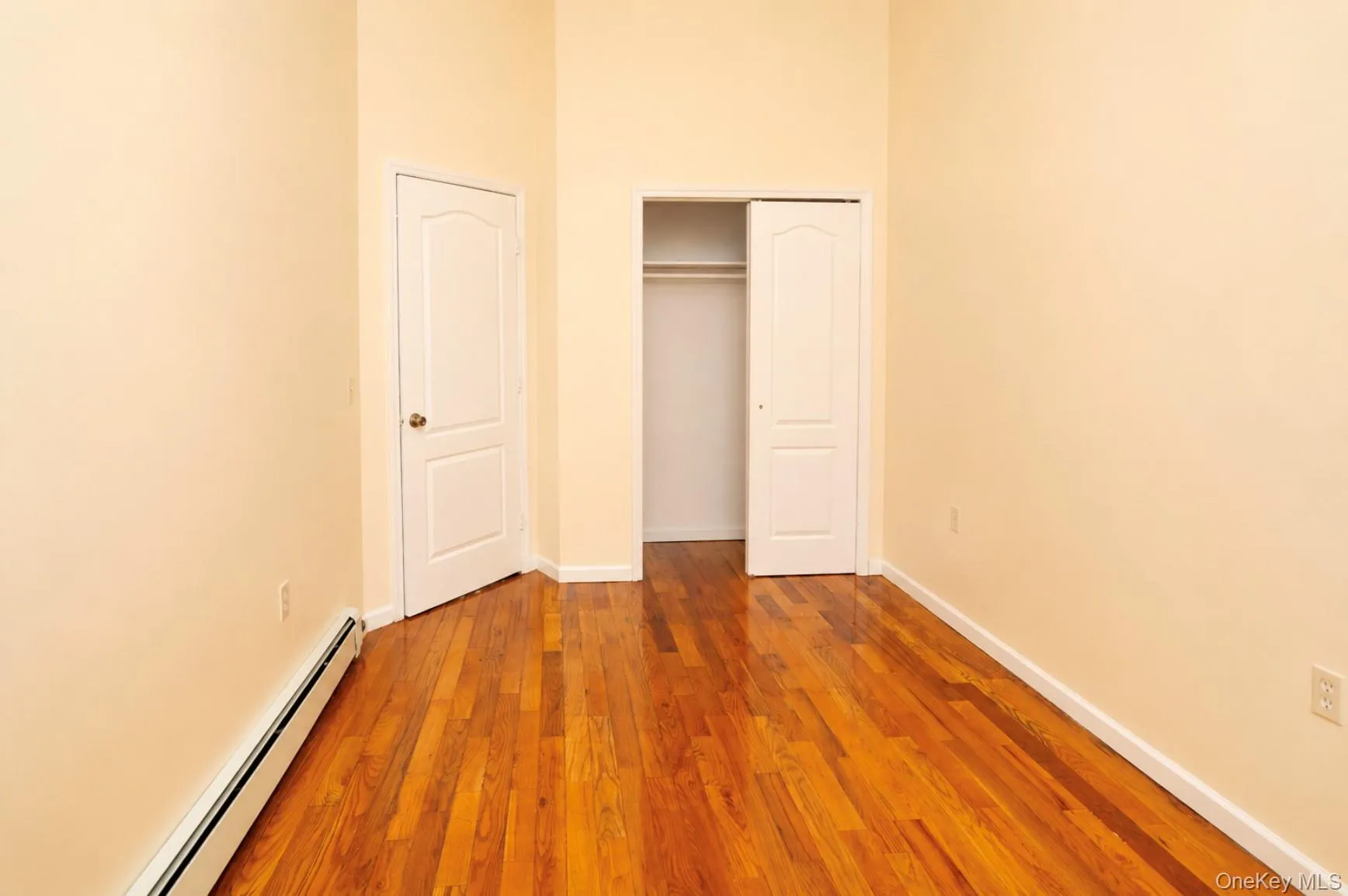 Spacious bedroom showcasing rich wood-finish flooring, soft neutral wall tones, and dual closet storage with white panel doors. Spacious bedroom showcasing rich wood-finish flooring, soft neutral wall tones, and dual closet storage with white panel doors.