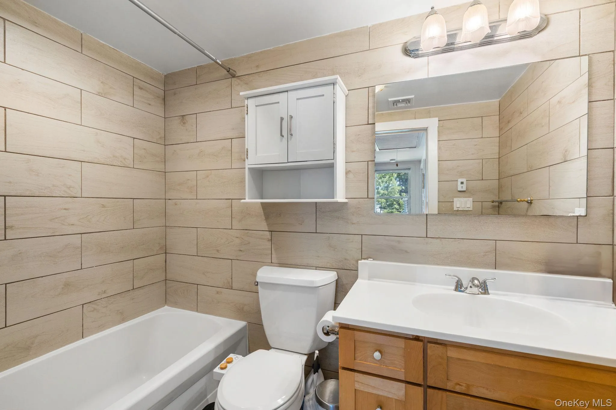 White ceramic bathtub, light wood-finish wall tiling, white toilet, single vanity with wood cabinetry, and a rectangular mirror White ceramic bathtub, light wood-finish wall tiling, white toilet, single vanity with wood cabinetry, and a rectangular mirror