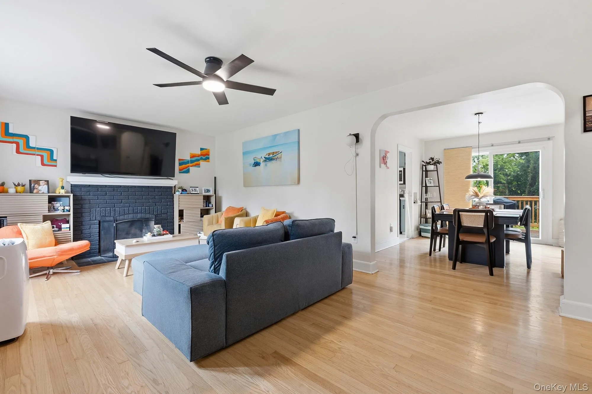 Open-concept living space featuring wood-finish flooring, a contemporary ceiling fan, and a painted brick fireplace Open-concept living space featuring wood-finish flooring, a contemporary ceiling fan, and a painted brick fireplace