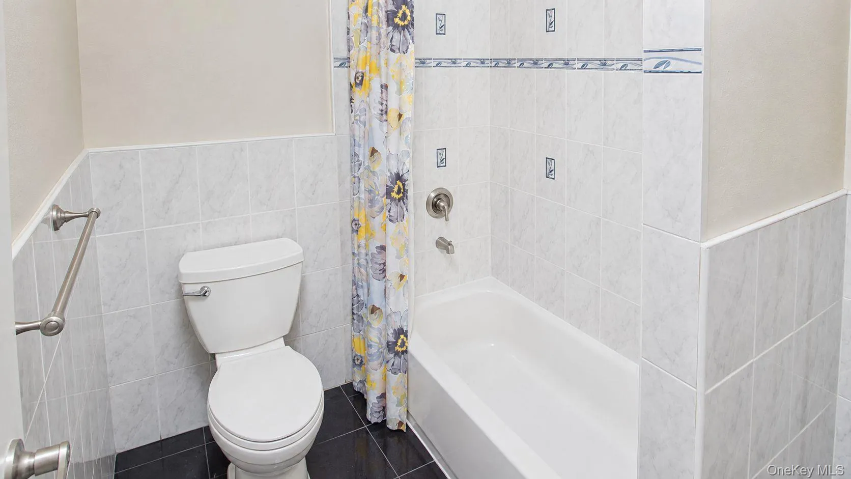 Bathroom featuring a white toilet, white bathtub with a chrome faucet, and light-colored wall tiling with decorative accents Bathroom featuring a white toilet, white bathtub with a chrome faucet, and light-colored wall tiling with decorative accents