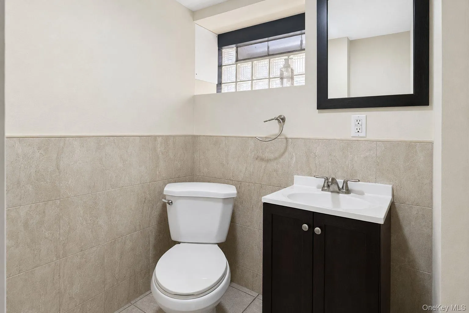 Bathroom featuring a dark wood-finish vanity with an integrated white sink, tiled walls, a framed mirror, and a glass block window Bathroom featuring a dark wood-finish vanity with an integrated white sink, tiled walls, a framed mirror, and a glass block window