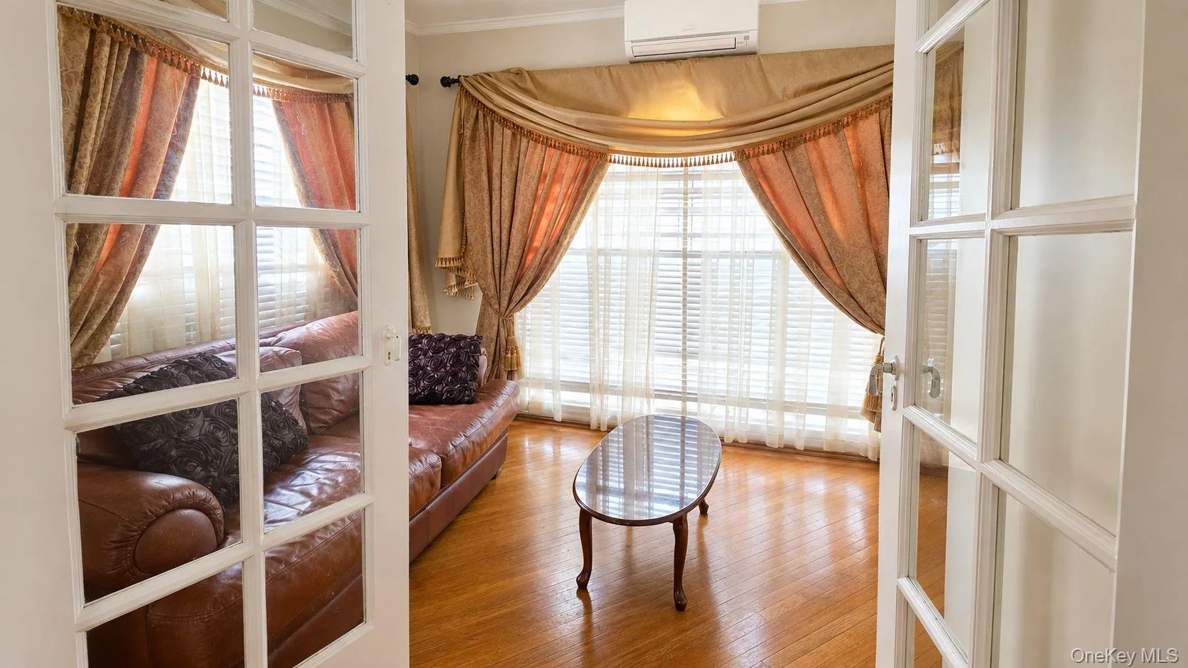 French doors opening to a living area featuring wood-finish flooring, a large window with horizontal blinds, and crown molding French doors opening to a living area featuring wood-finish flooring, a large window with horizontal blinds, and crown molding
