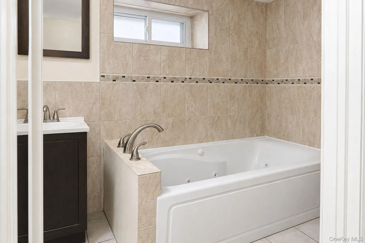 White jetted soaking tub with deck-mounted brushed nickel fixtures, surrounded by beige stone-look wall tiles White jetted soaking tub with deck-mounted brushed nickel fixtures, surrounded by beige stone-look wall tiles