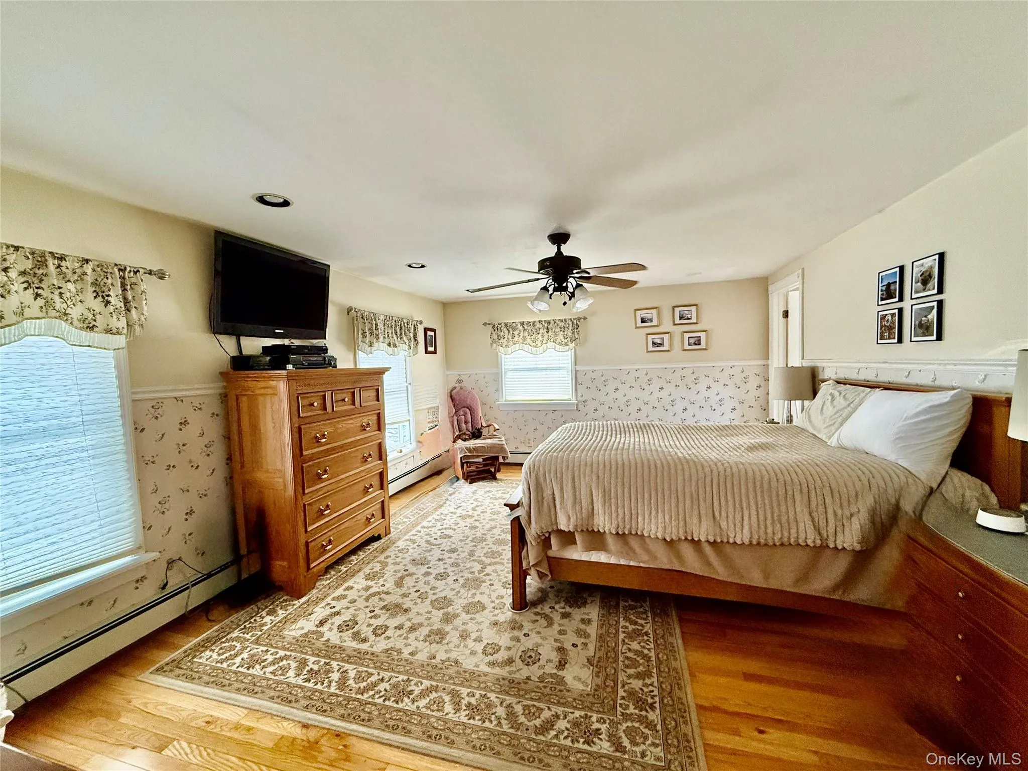 Bedroom featuring a baseboard heating unit, a wainscoted wall, a ceiling fan, light wood-style flooring, and wallpapered walls Bedroom featuring a baseboard heating unit, a wainscoted wall, a ceiling fan, light wood-style flooring, and wallpapered walls