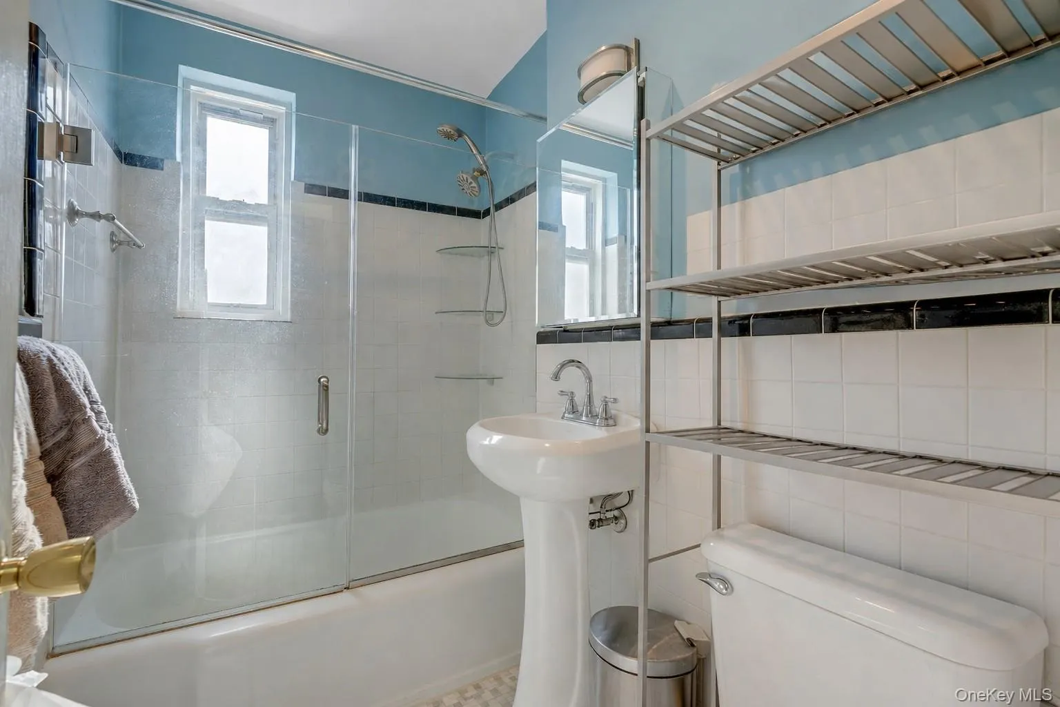 Bathroom featuring a pedestal sink, white tile walls with a black accent band, a tub with a glass sliding shower door, a mounted shower head, and a window Bathroom featuring a pedestal sink, white tile walls with a black accent band, a tub with a glass sliding shower door, a mounted shower head, and a window
