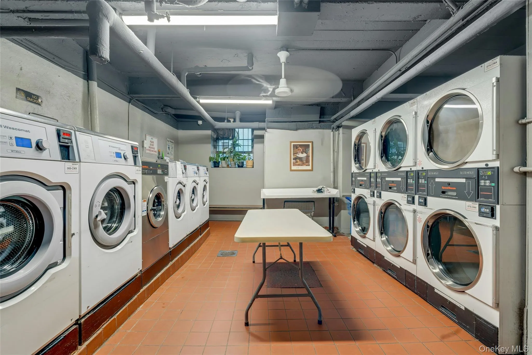 On-site laundry facility featuring multiple front-loading washers and dryers, terracotta-style flooring, a window providing natural light, exposed piping, and ceiling-mounted lighting On-site laundry facility featuring multiple front-loading washers and dryers, terracotta-style flooring, a window providing natural light, exposed piping, and ceiling-mounted lighting