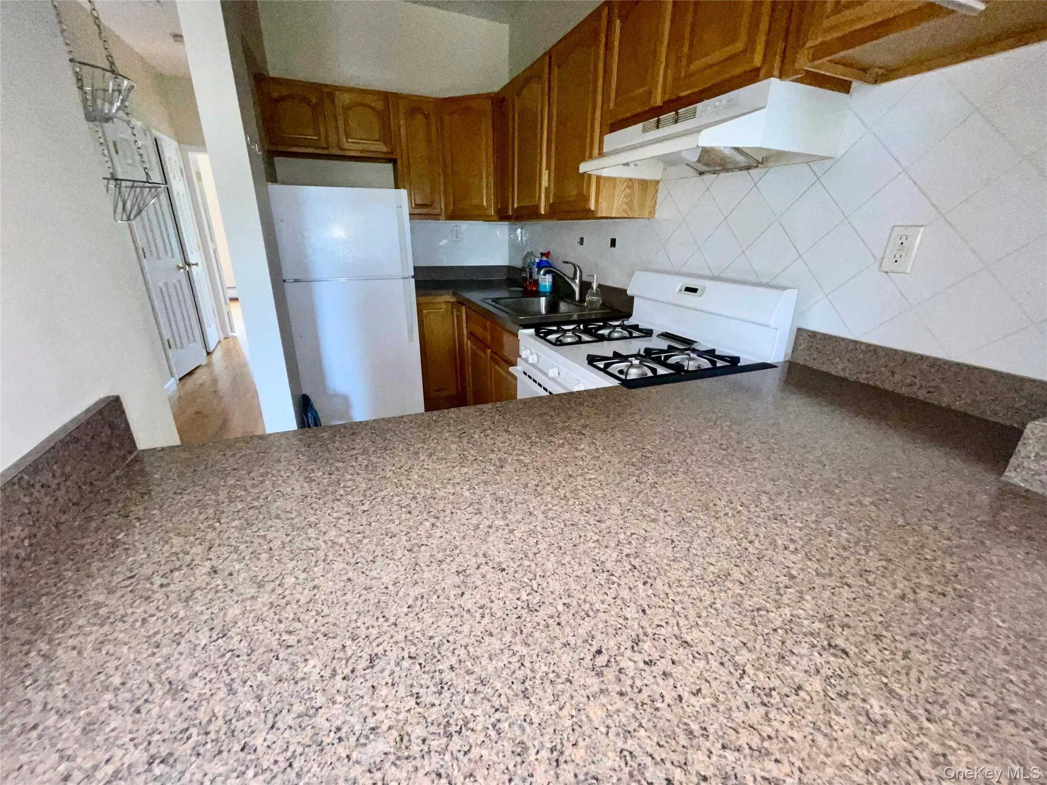 Kitchen with white appliances, under cabinet range hood, brown cabinetry, tasteful backsplash, and dark countertops Kitchen with white appliances, under cabinet range hood, brown cabinetry, tasteful backsplash, and dark countertops