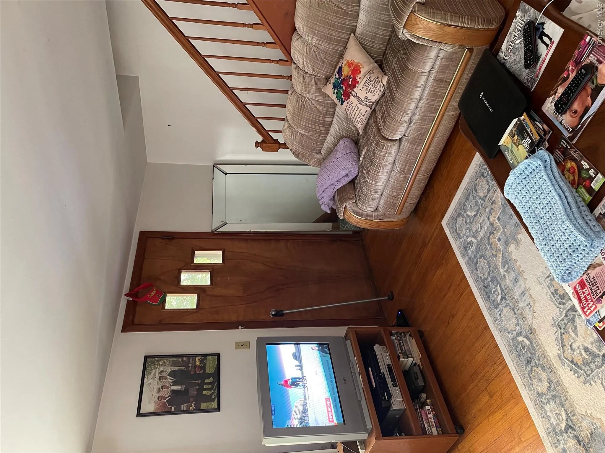 Comfortable living room featuring hardwood flooring, a striped upholstered sofa with rolled arms, wooden staircase with metal railings, and a wood-finish entry door with decorative candle sconces Comfortable living room featuring hardwood flooring, a striped upholstered sofa with rolled arms, wooden staircase with metal railings, and a wood-finish entry door with decorative candle sconces