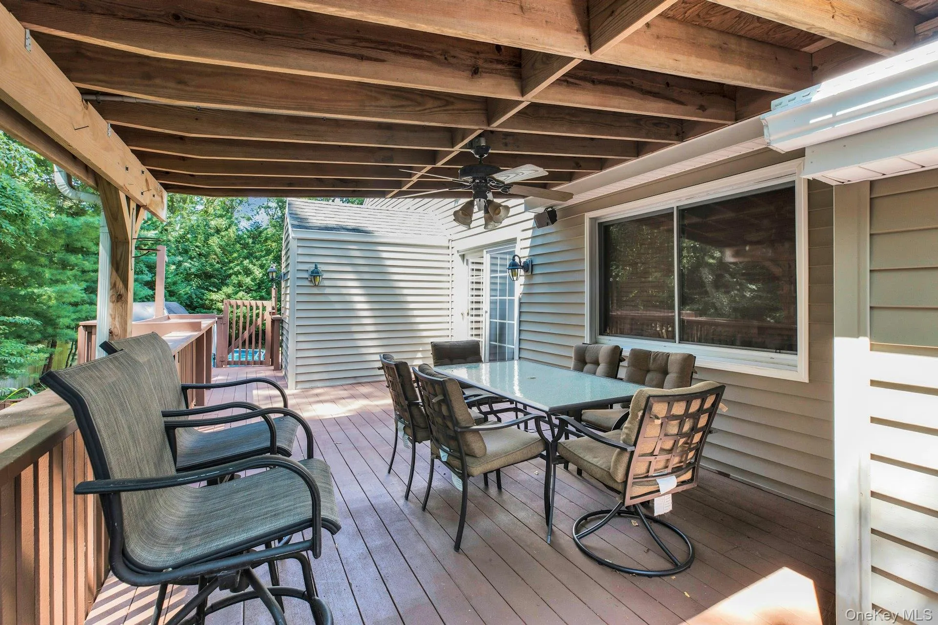 Expansive covered deck featuring wood-finish flooring, exposed wood beams, a ceiling fan, and direct access to interior spaces through a sliding glass door Expansive covered deck featuring wood-finish flooring, exposed wood beams, a ceiling fan, and direct access to interior spaces through a sliding glass door