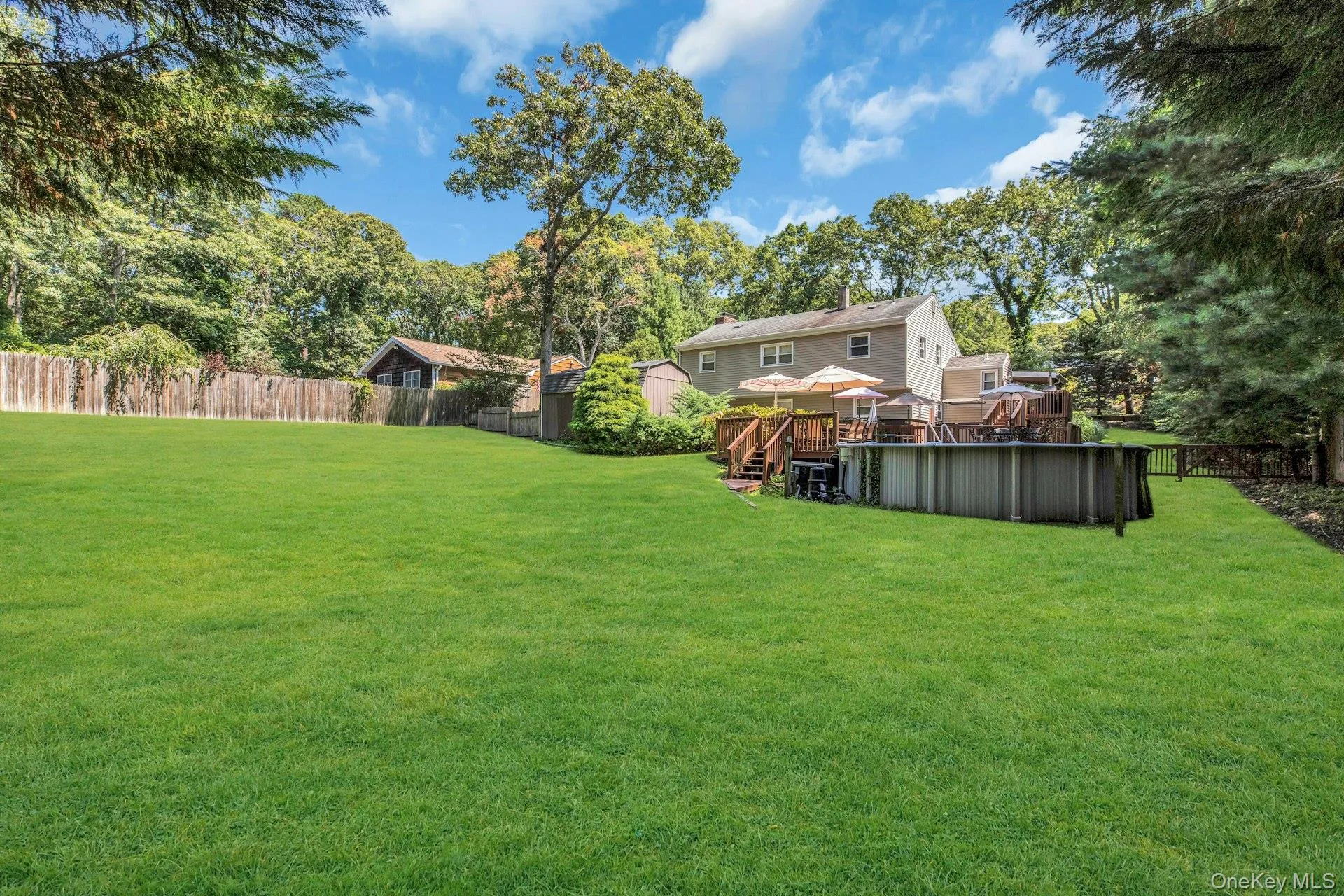Expansive manicured lawn with mature tree canopy, wooden deck with integrated seating, detached storage structure, and fenced perimeter Expansive manicured lawn with mature tree canopy, wooden deck with integrated seating, detached storage structure, and fenced perimeter