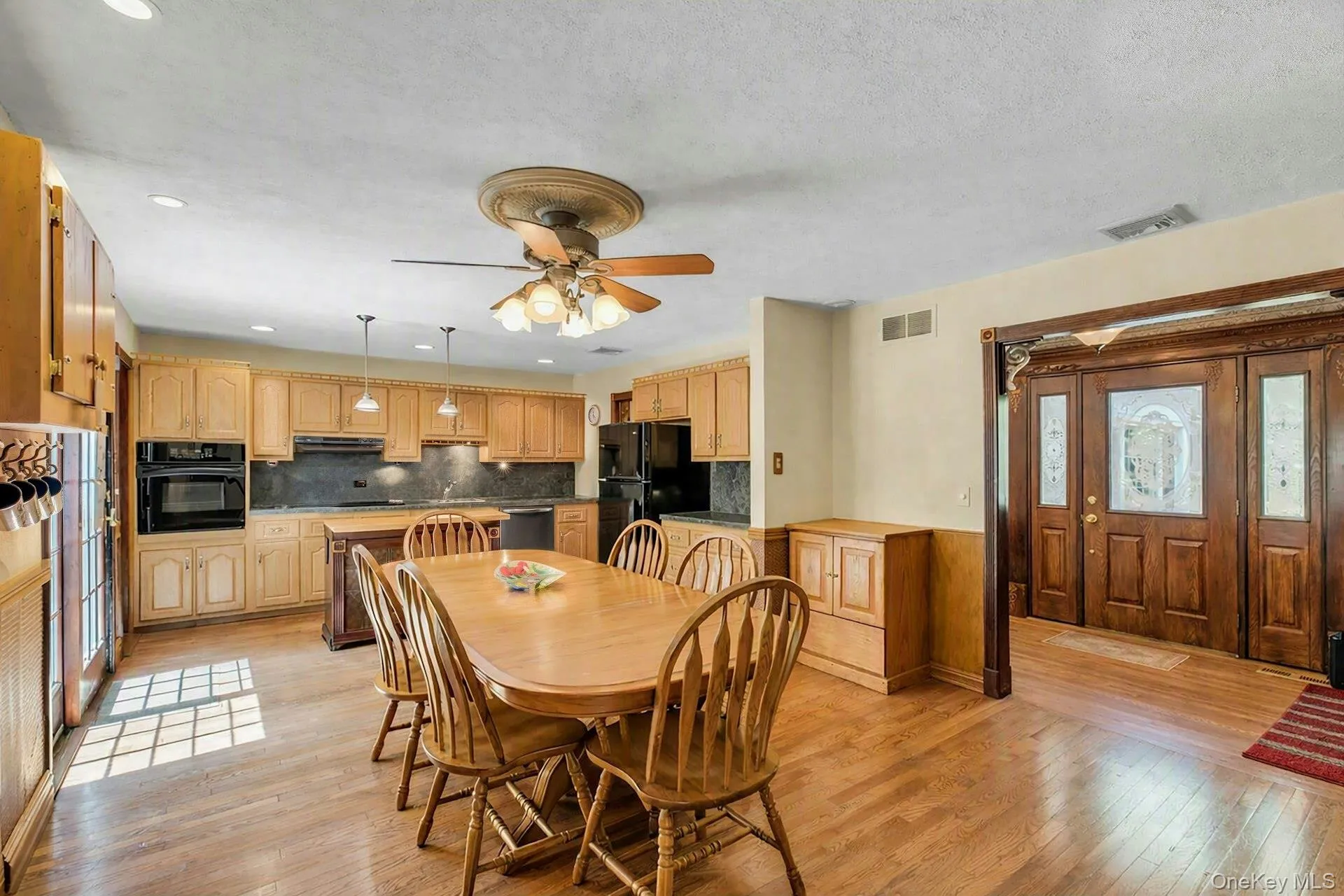 Spacious kitchen and dining area featuring warm wood cabinetry, stainless steel appliances, wood-finish flooring throughout, a ceiling fan with integrated lighting, and a generous dining table with seating for six Spacious kitchen and dining area featuring warm wood cabinetry, stainless steel appliances, wood-finish flooring throughout, a ceiling fan with integrated lighting, and a generous dining table with seating for six