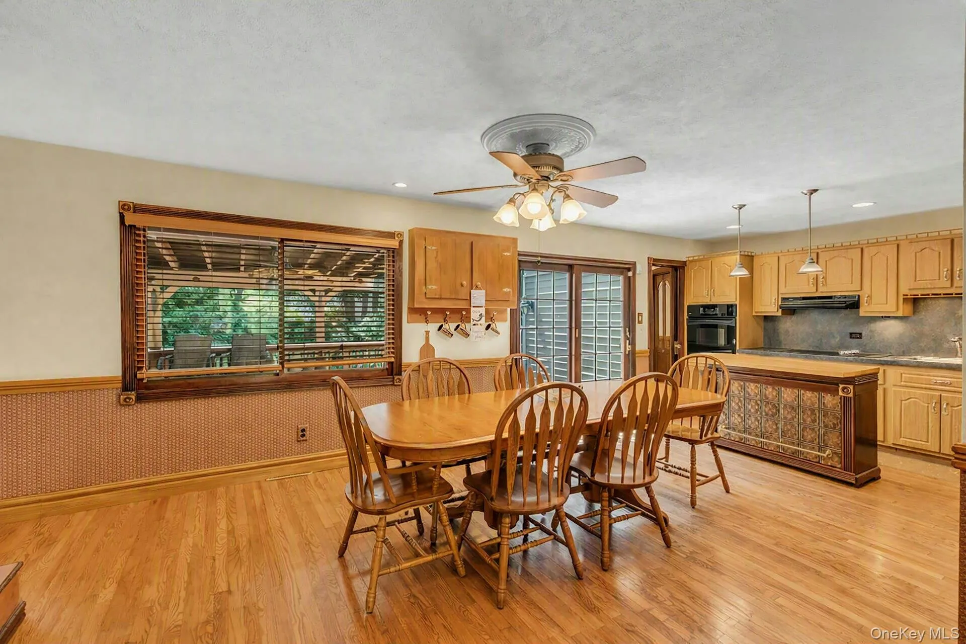 Spacious open-concept dining and kitchen area featuring warm wood-finish flooring throughout, a round pedestal dining table with seating for four, custom wood cabinetry with ample storage, stainless steel appliances, pendant lighting over the kitchen island, and a ceiling fan with integrated light fixture Spacious open-concept dining and kitchen area featuring warm wood-finish flooring throughout, a round pedestal dining table with seating for four, custom wood cabinetry with ample storage, stainless steel appliances, pendant lighting over the kitchen island, and a ceiling fan with integrated light fixture