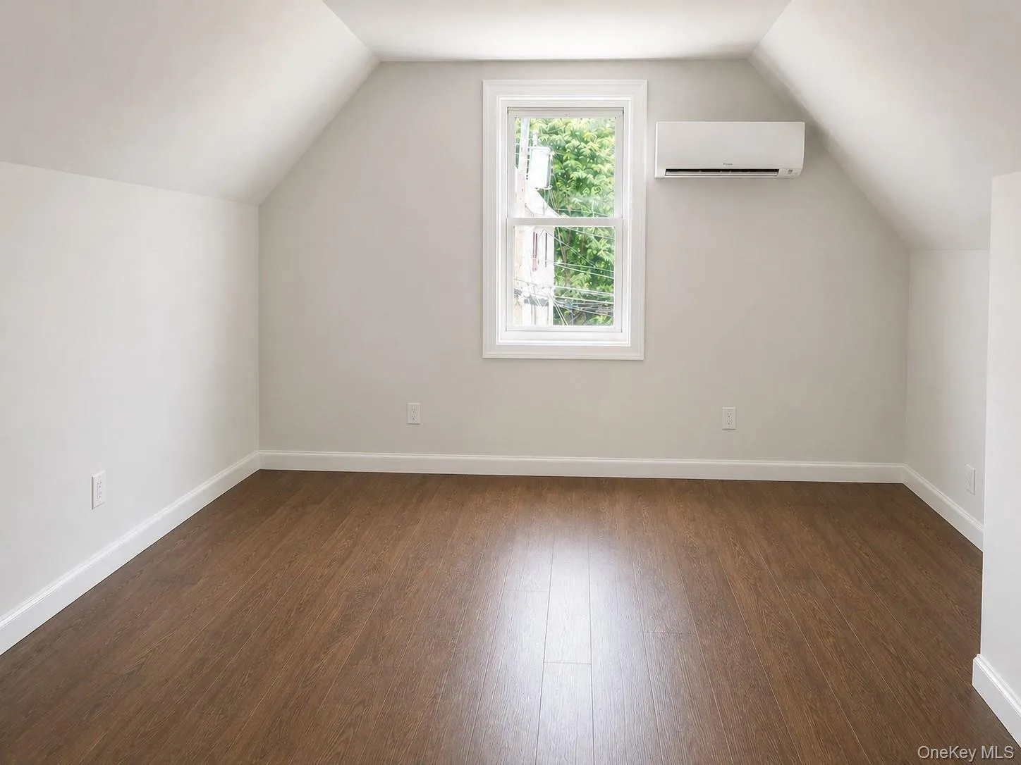 Versatile room featuring wood-finish flooring, crisp white baseboards, a white window frame, and an installed wall-mounted air conditioning unit Versatile room featuring wood-finish flooring, crisp white baseboards, a white window frame, and an installed wall-mounted air conditioning unit
