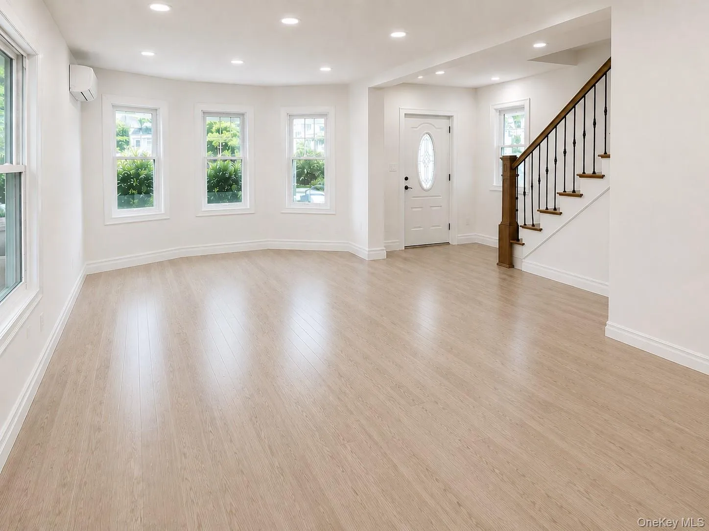 Open-concept living area featuring light wood-finish flooring, recessed lighting, and a prominent staircase with dark wood newel post and wrought iron balusters Open-concept living area featuring light wood-finish flooring, recessed lighting, and a prominent staircase with dark wood newel post and wrought iron balusters
