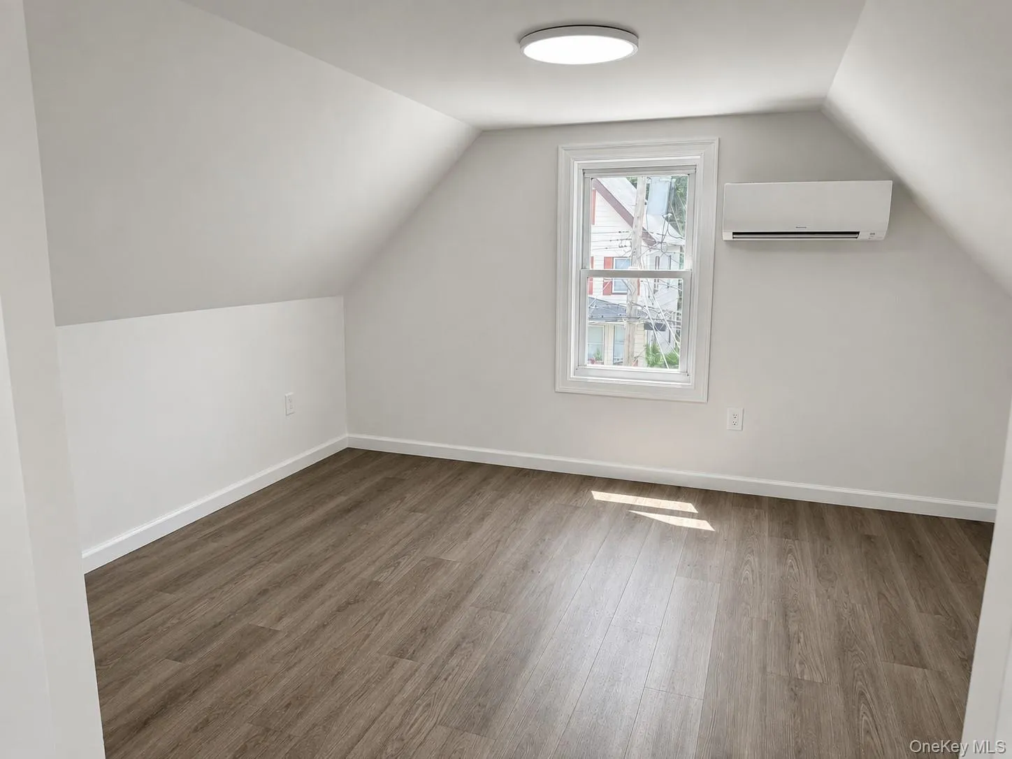 Attic space featuring wood-finish flooring, a white-framed window, and a wall-mounted climate control unit Attic space featuring wood-finish flooring, a white-framed window, and a wall-mounted climate control unit