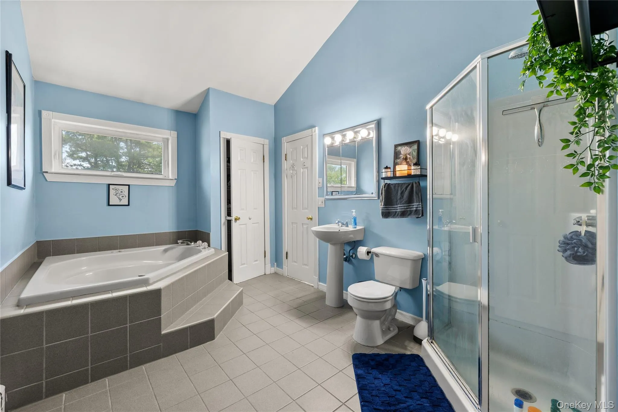 Spacious primary and bathroom featuring a built-in soaking tub with tiled surround, a separate glass-enclosed shower, pedestal sink, and a window providing natural light Dual entrance to hall Spacious primary and bathroom featuring a built-in soaking tub with tiled surround, a separate glass-enclosed shower, pedestal sink, and a window providing natural light Dual entrance to hall